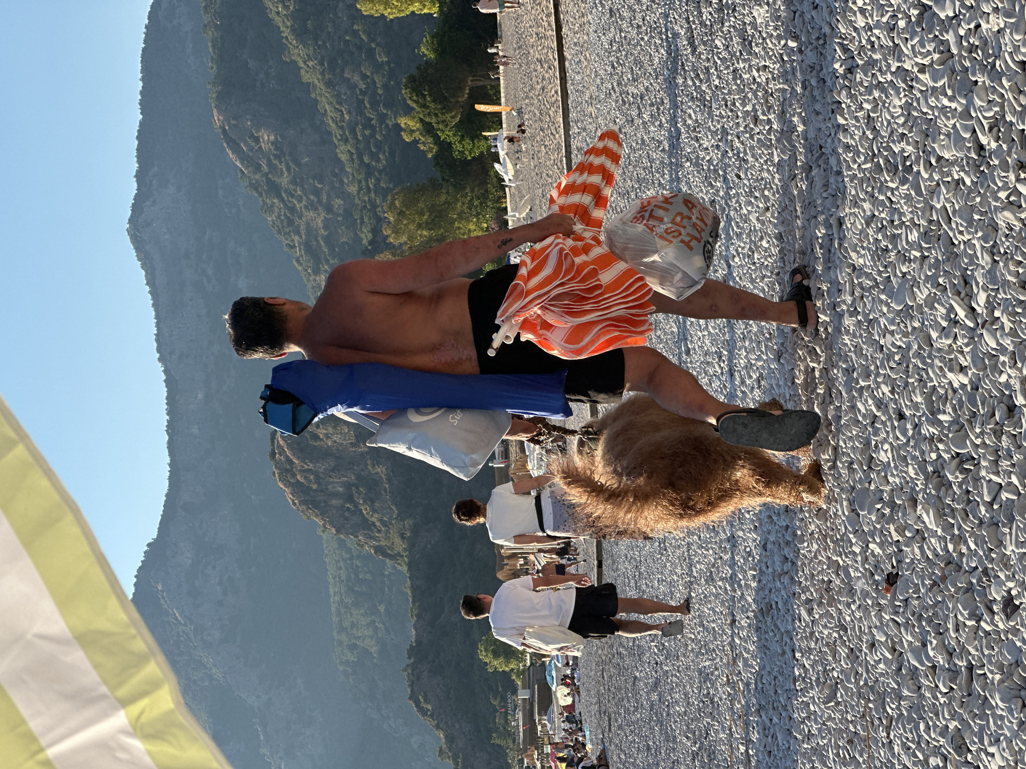 This media file captures a sunny, late afternoon scene on a pebble beach, likely in Fethiye, Turkey, given the context. In the foreground, a shirtless man with dark hair is walking away from the camera, carrying several beach items. He has a blue beach chair or mat folded over his left shoulder and an orange and white striped beach umbrella in his right hand, along with a transparent plastic bag.
Beside him, a shaggy, light brown dog with curly fur is walking on the pebbles. The beach itself is covered in countless smooth, light-colored, oval-shaped stones, casting long shadows to the left, indicating the late afternoon sun. In the mid-ground, other people are visible on the beach; one man in a white t-shirt and dark shorts is walking with a white bag, and another person further back is also seen.
The background is dominated by large, green-covered mountains rising steeply under a clear blue sky. These mountains show rugged textures and are densely vegetated. A small, faint tattoo is visible on the shirtless man's left upper arm.
Visible text includes partial Turkish words on the plastic bag, which appear to include "ATIK" (meaning "waste") and "HAYAT" (meaning "life"). On a grey bag carried by a person in the mid-ground, the text "JAM300" is discernible. The overall atmosphere suggests a leisurely day at the beach, transitioning into the evening.