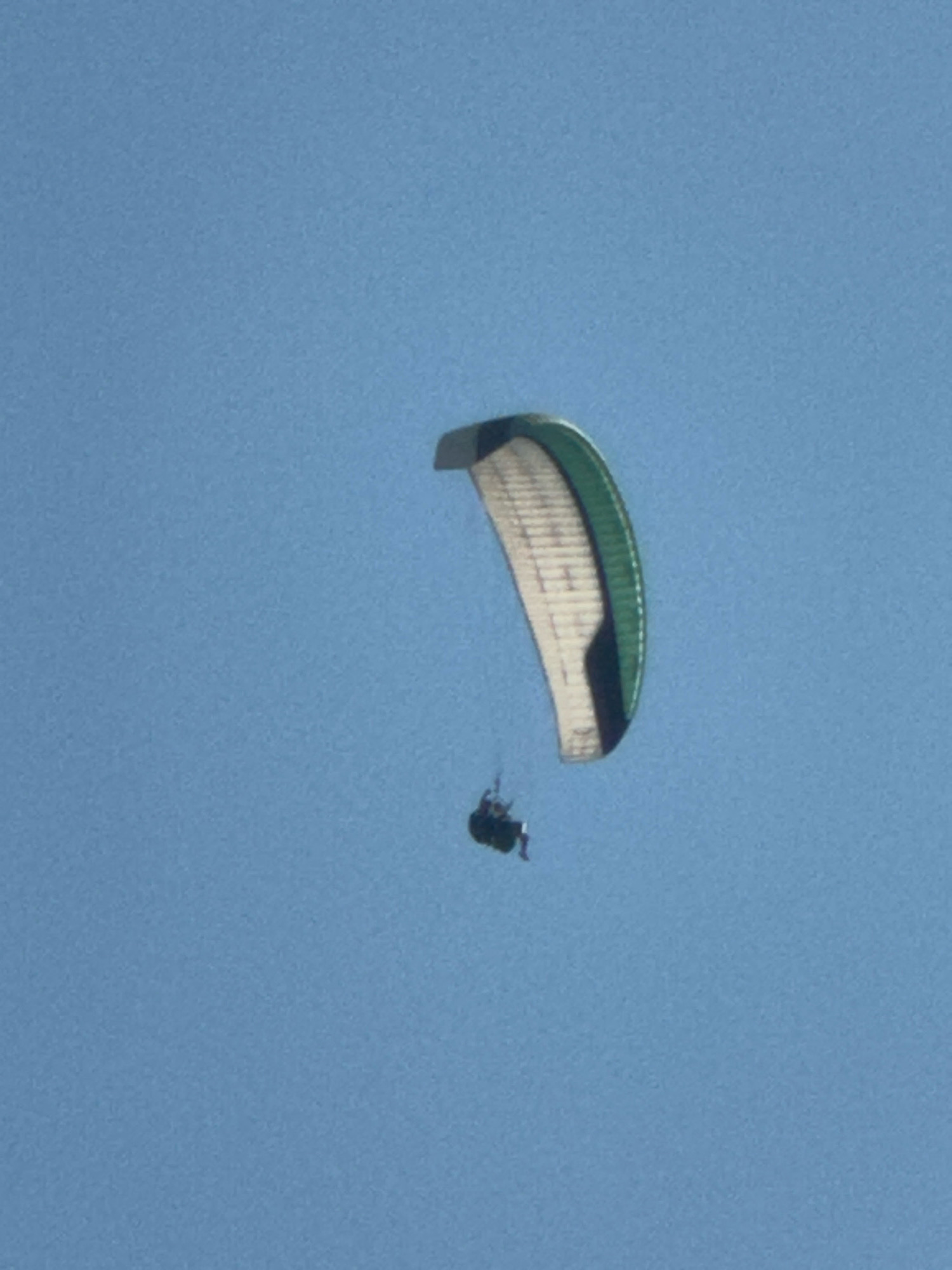 In the city of Boreti, Montenegro, a paraglider is captured mid-flight against a clear blue sky. The paraglider's canopy, predominantly white with green and black accents, is inflated and arching gracefully. Suspended beneath the canopy, a person is seated in a harness, their legs extended. The individual appears to be piloting the paraglider, engaged in the activity of paragliding. The environment is an open sky, suggesting an outdoor recreational activity.