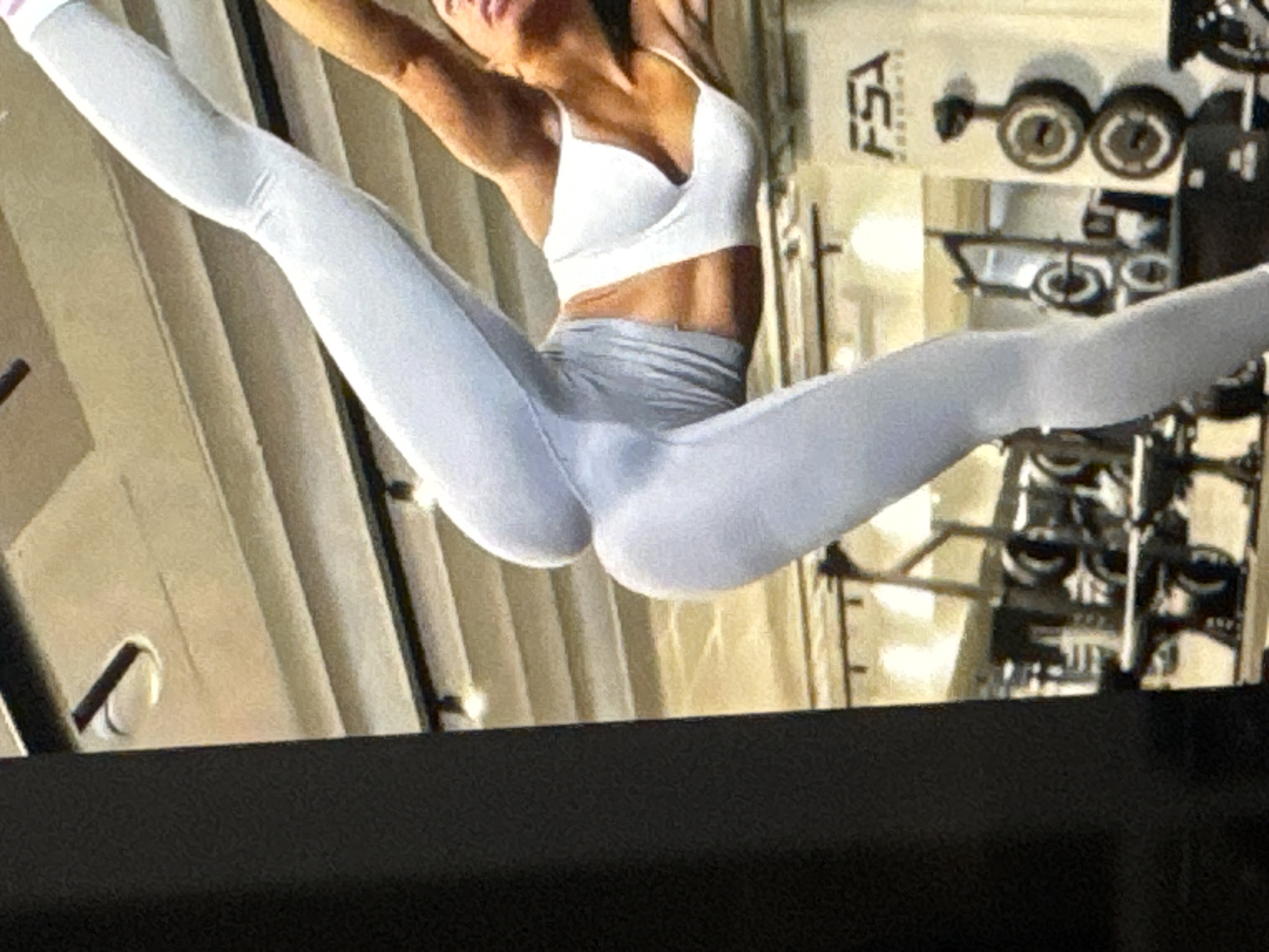 An athletic woman is performing a yoga pose in a gym. She is wearing a light grey sports bra and matching leggings. Her body is contorted, with one leg extended upwards and the other bent. The gym appears to be well-equipped, with various weight machines and other fitness equipment visible in the background. The environment is dimly lit with overhead lights illuminating the space. The woman's physique suggests she is physically fit and dedicated to her training. The gym itself seems to be a professional establishment catering to fitness enthusiasts.