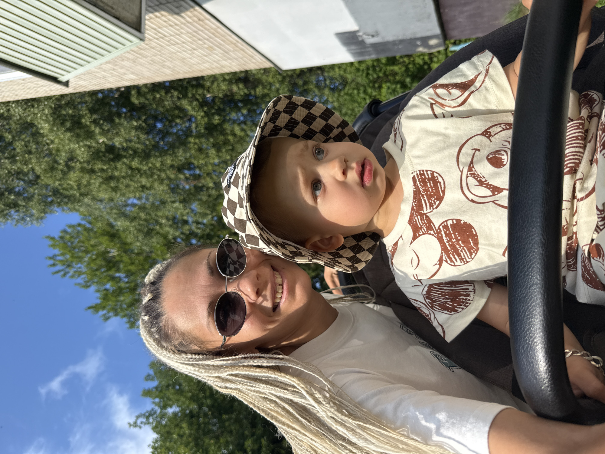 In Rusanovo, Russia, under a bright blue sky with scattered clouds, a woman with blonde braided hair and sunglasses smiles at the camera, her left arm resting on a stroller. Next to her, a young child wearing a checkered bucket hat and a light-colored t-shirt with a brown graphic print looks off to the right. They are positioned outdoors, with a building and lush green trees in the background. The scene appears to be captured on a sunny day, likely during daylight hours.