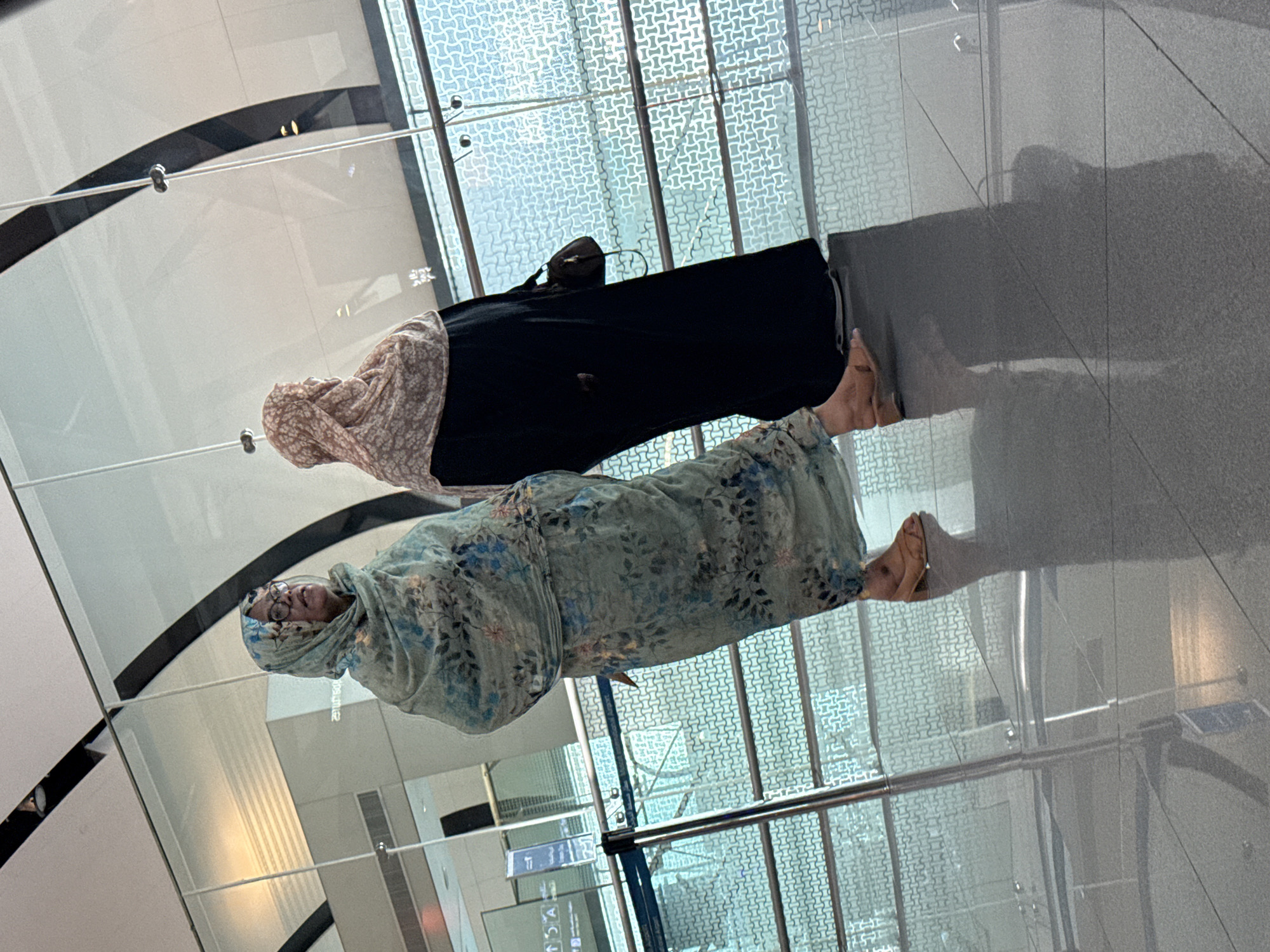 In Seeb Province, Oman, two women are standing in an airport terminal. The woman on the left wears a light blue hijab with a floral pattern and glasses. She is wearing a matching patterned dress and brown sandals. The woman on the right wears a light pink hijab with a floral pattern and a black abaya. She is carrying a dark brown handbag. They are positioned in front of a large glass window that overlooks a patterned walkway or exterior area. The floor is highly reflective, showing the reflections of the women and the terminal environment.