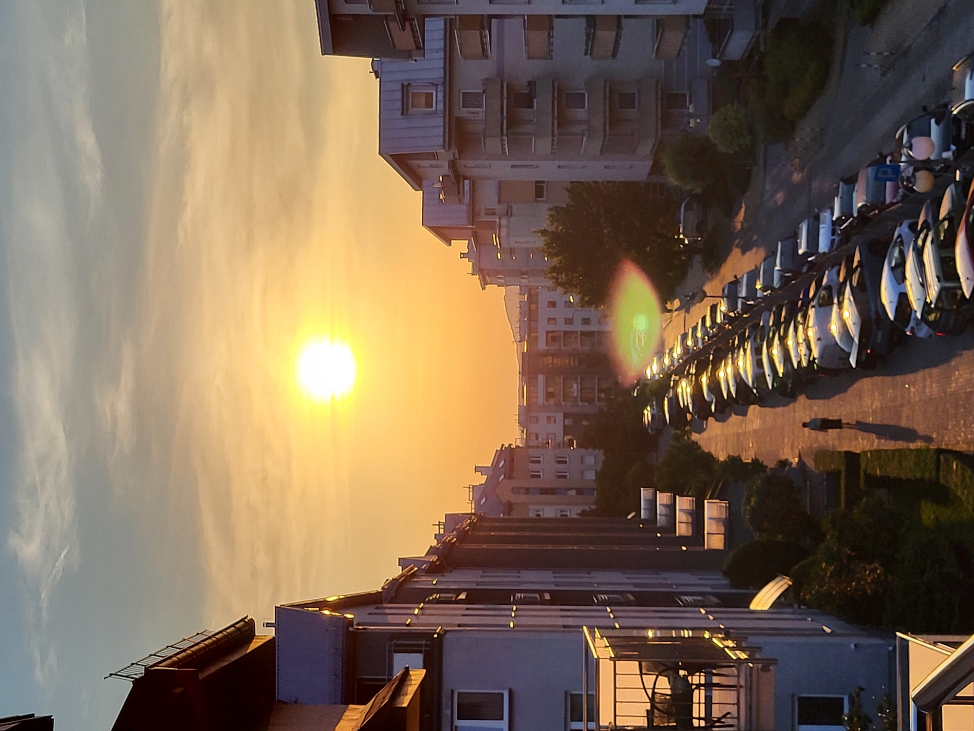 The image depicts a street lined with apartment buildings and parked cars under a warm, setting sun. The sun is prominent in the sky, casting a golden hue over the scene and creating long shadows. A single person is walking down the sidewalk. The buildings are multi-story residential structures with balconies and windows. The street is paved and appears to be in a residential area of a city. The overall impression is of a tranquil evening in an urban environment.