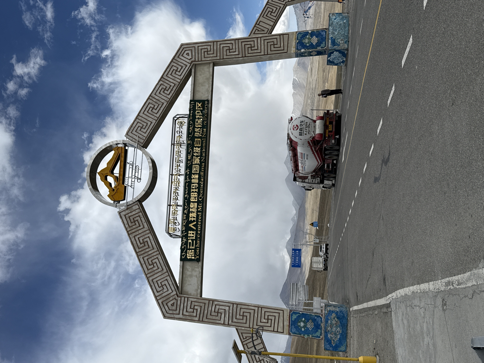 This daytime image captures the entrance to a protected area, likely a checkpoint, in Lhatsé, China. A prominent, ornate archway with a repeating geometric pattern spans a paved road. Central to the archway is a circular emblem and a large sign in Tibetan, Chinese, and English. The English text clearly states: "You have entered Mt. Qomolangma National Natre Reserve," identifying the location as the Mount Everest (Qomolangma) National Nature Reserve.
On the right side of the road, a large white and red tanker truck, bearing the Chinese characters "杨嘉汽车" (Yang Jia Automobile) and the number "AF9008," is visible. A person in dark attire stands near the truck. The road features white dashed lines and a solid yellow line, with a smaller blue sign in the distance showing Chinese text "十次超速十次客" (Ten times speeding, ten times passenger), likely a traffic regulation for commercial vehicles. The backdrop features rugged, distant mountains under a sky with scattered clouds. Blue decorative panels adorn the bases of the archway's pillars.