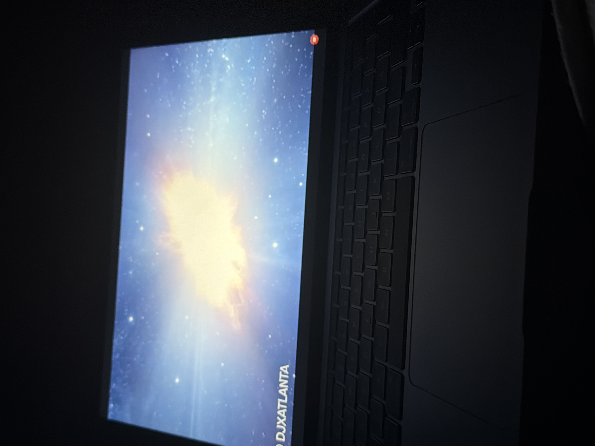 The image shows a laptop with a galaxy-themed wallpaper displayed on its screen. Text that reads "DJXATLANTA" is visible on the lower left portion of the wallpaper. The laptop is black and open, with its keyboard and trackpad visible. The lighting is dim, suggesting the photo was taken in a dark environment. The image does not contain information to determine its location within the city of Plovdiv, Bulgaria.