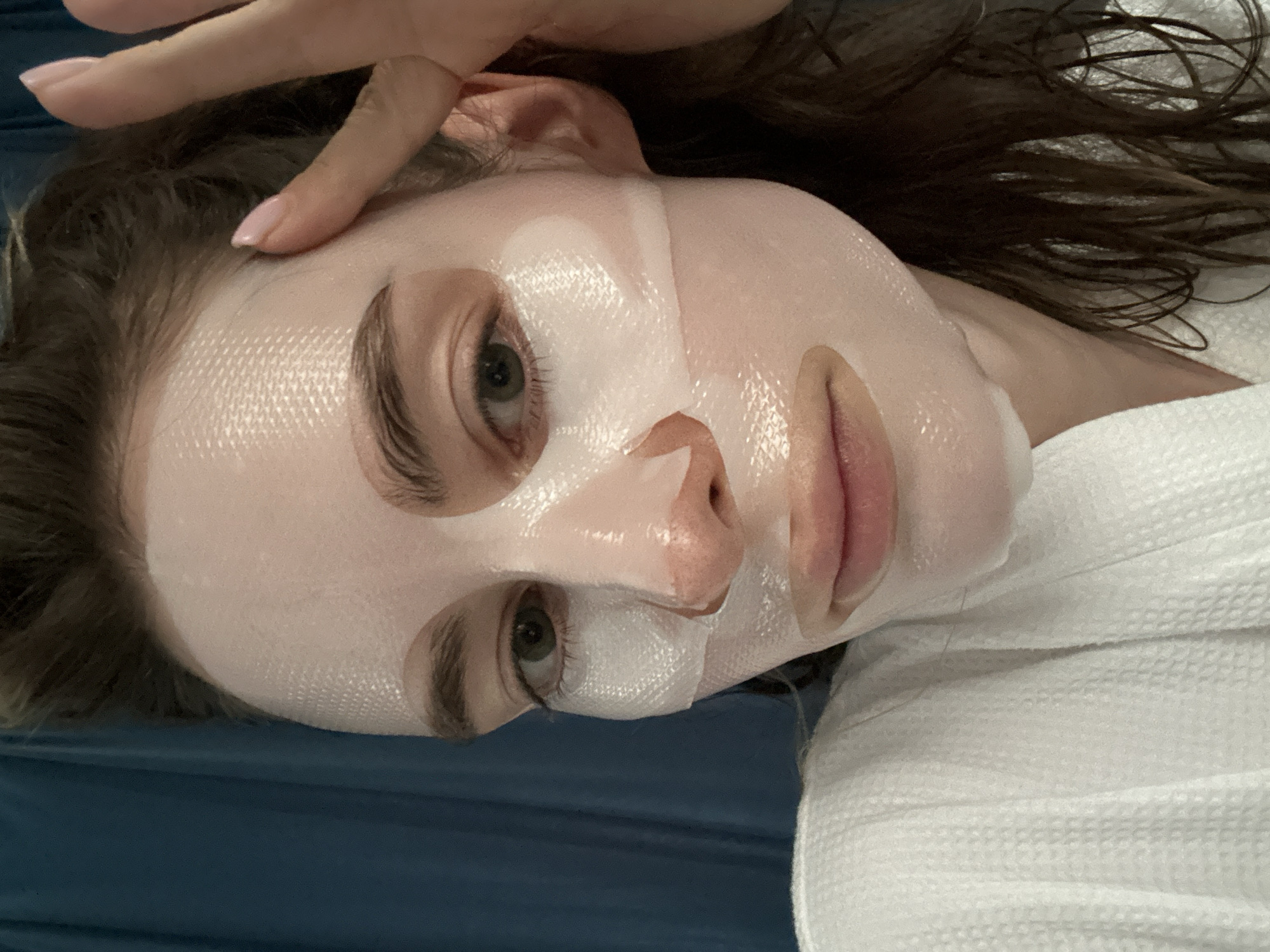 A female individual is depicted in a close-up, chest-up shot within an indoor setting in Choeng Thale, Thailand. She is wearing a white, textured sheet mask that covers her forehead, cheeks, and chin, with cutouts revealing her eyes, nose, and mouth. The mask exhibits a fine, grid-like pattern. Her left eye, visible, is green. Her right hand, with light pink fingernail polish, is raised with fingers touching the right side of her forehead, near her temple, above the mask. Her hair, dark brown and appearing damp, frames her face and shoulders. She wears a white, textured bathrobe, visible on her left shoulder and chest. The background consists of a dark blue, plain surface, consistent with a curtain.
