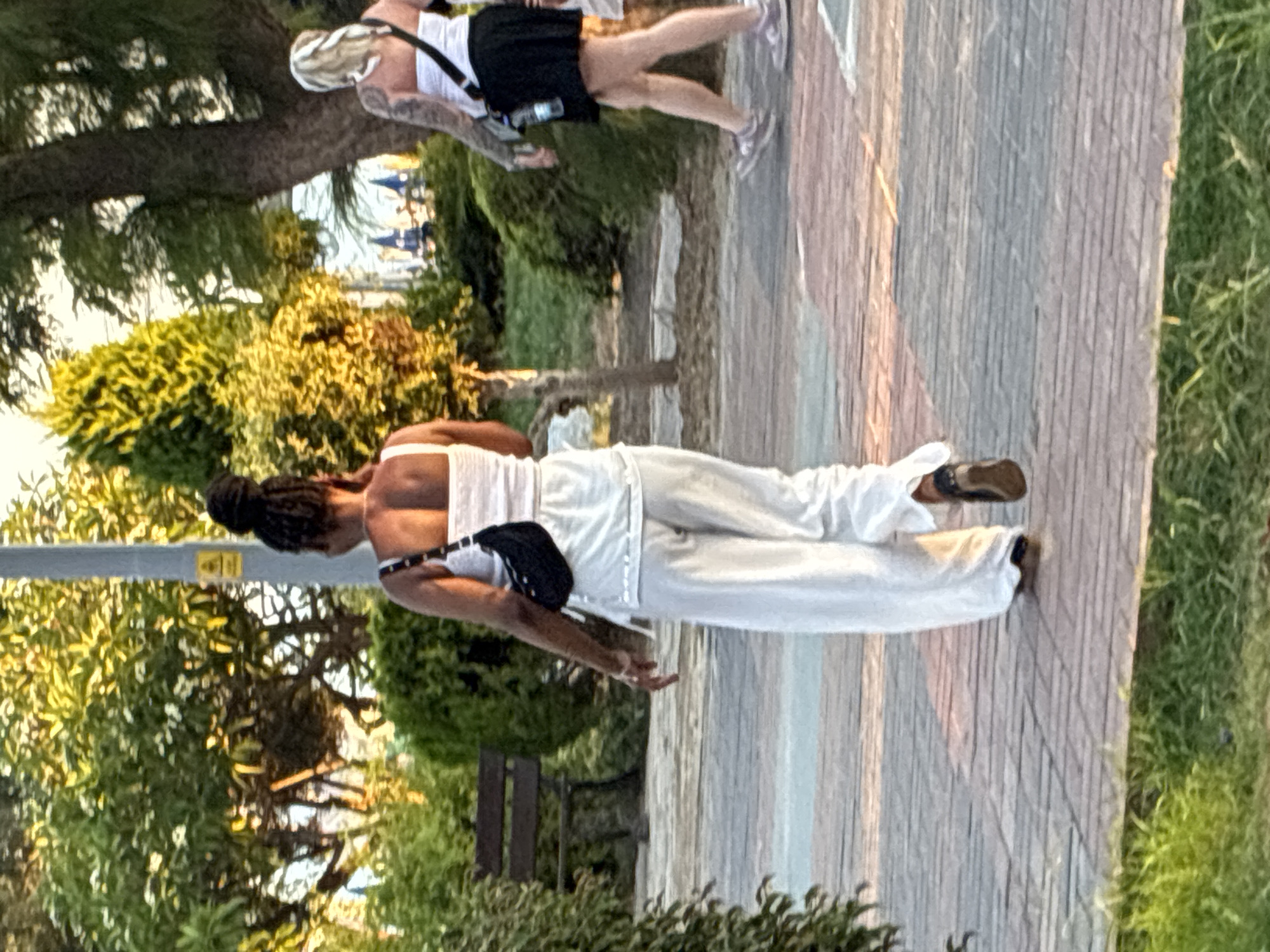 The image captures a woman walking away from the camera on a paved walkway in a park-like setting in Alanya, Turkey. She is wearing white, loose-fitting pants and a white top, with a black crossbody bag. The sun is setting, casting a warm glow on the scene. In the background, there is lush greenery, trees, a park bench, and another woman walking in the distance. The walkway is made of interlocking paving stones.