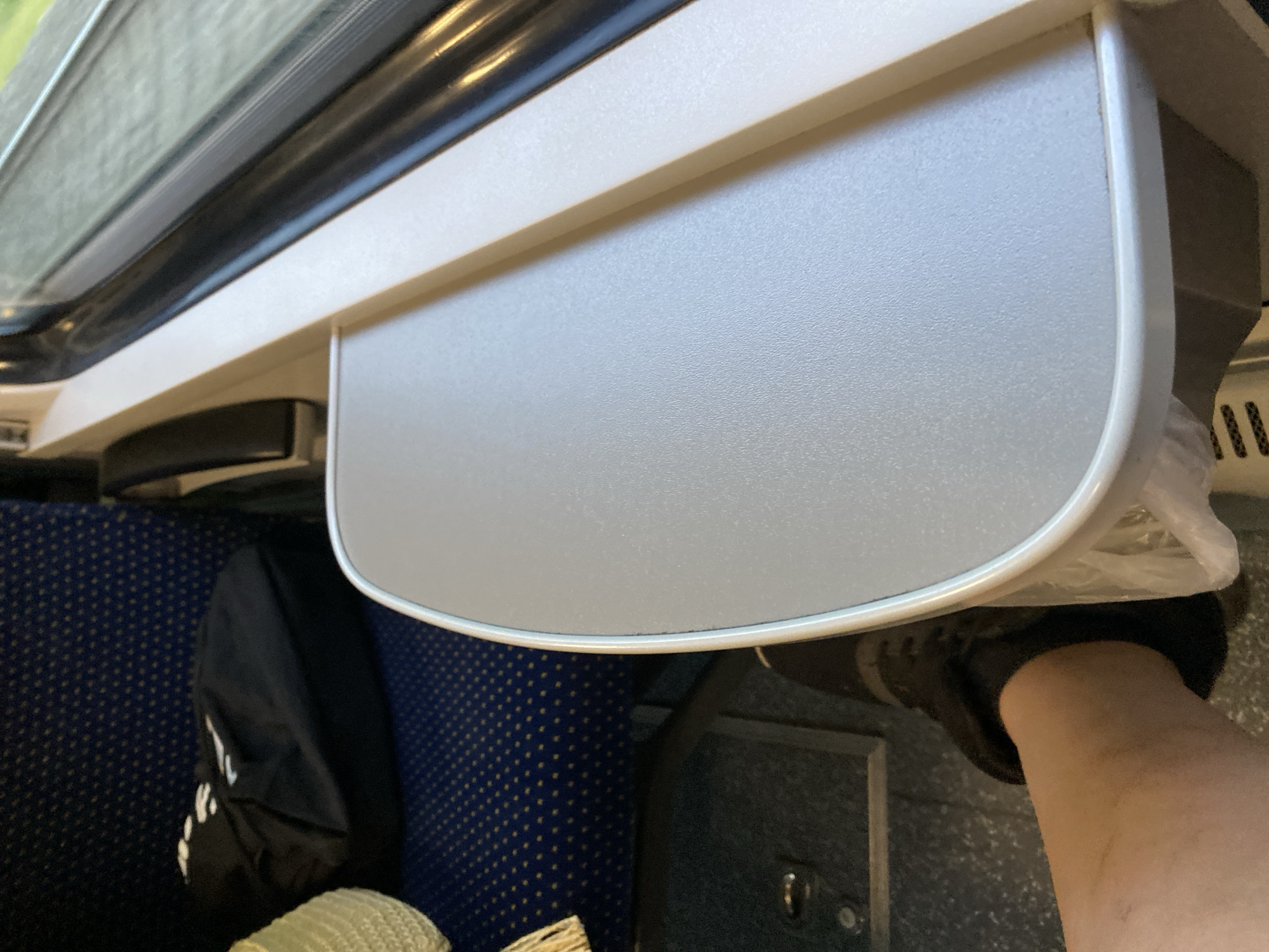 The image depicts a close-up view of a tray table on a train, likely in Bílina, Czechia. The tray table is light gray and positioned at an angle, with a beige edge along its length. To the left of the tray, a portion of a seat with a blue and yellow patterned fabric is visible. A black bag rests on the seat. In the foreground, a person's leg and foot wearing a black shoe are visible, positioned beneath the tray table. The background shows a window with a view of what appears to be greenery. The overall scene suggests a passenger's perspective from a train seat.