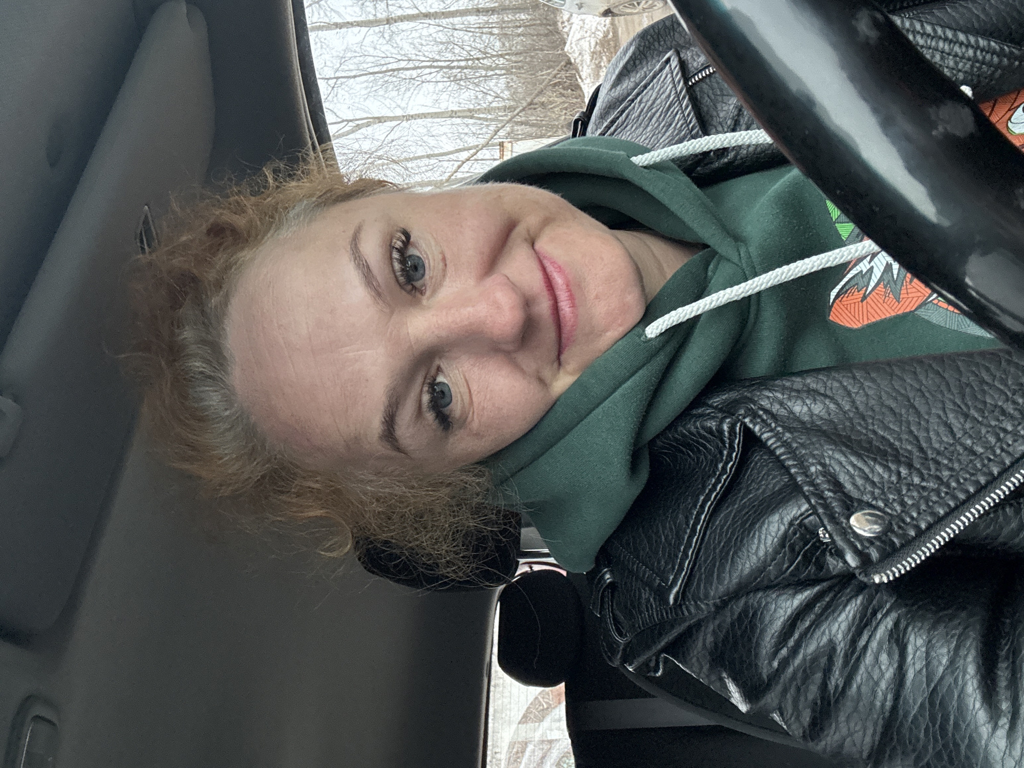 A woman with fair skin and light red hair, wearing a green hooded sweatshirt and a black leather jacket, is sitting in the driver's seat of a car. The car's interior is visible, including the steering wheel and the sun visor. Through the windshield, bare trees and possibly a snowy or overcast sky can be seen. Based on the location metadata, this scene is in Chaikovsky, Russia.