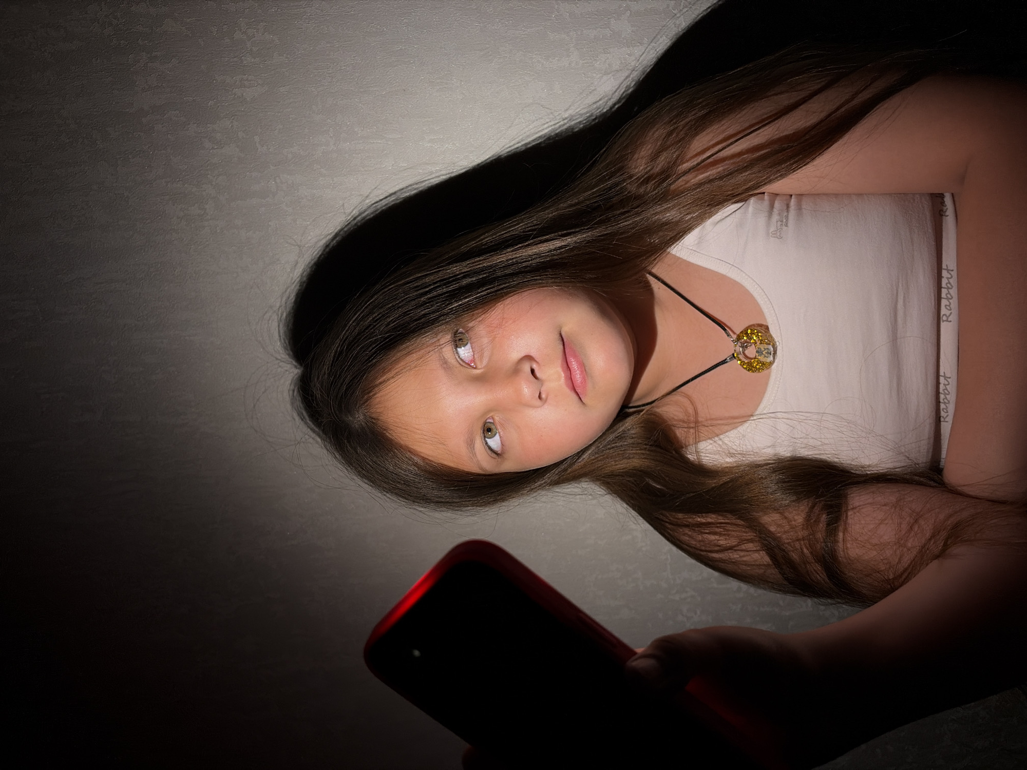 A young girl with long brown hair is looking at a red mobile phone screen in the dark. She is wearing a white tank top and a necklace with a round, yellow pendant. The background is a textured gray wall. The image is dimly lit, casting shadows on the wall behind the girl. The location is not discernable from the image.