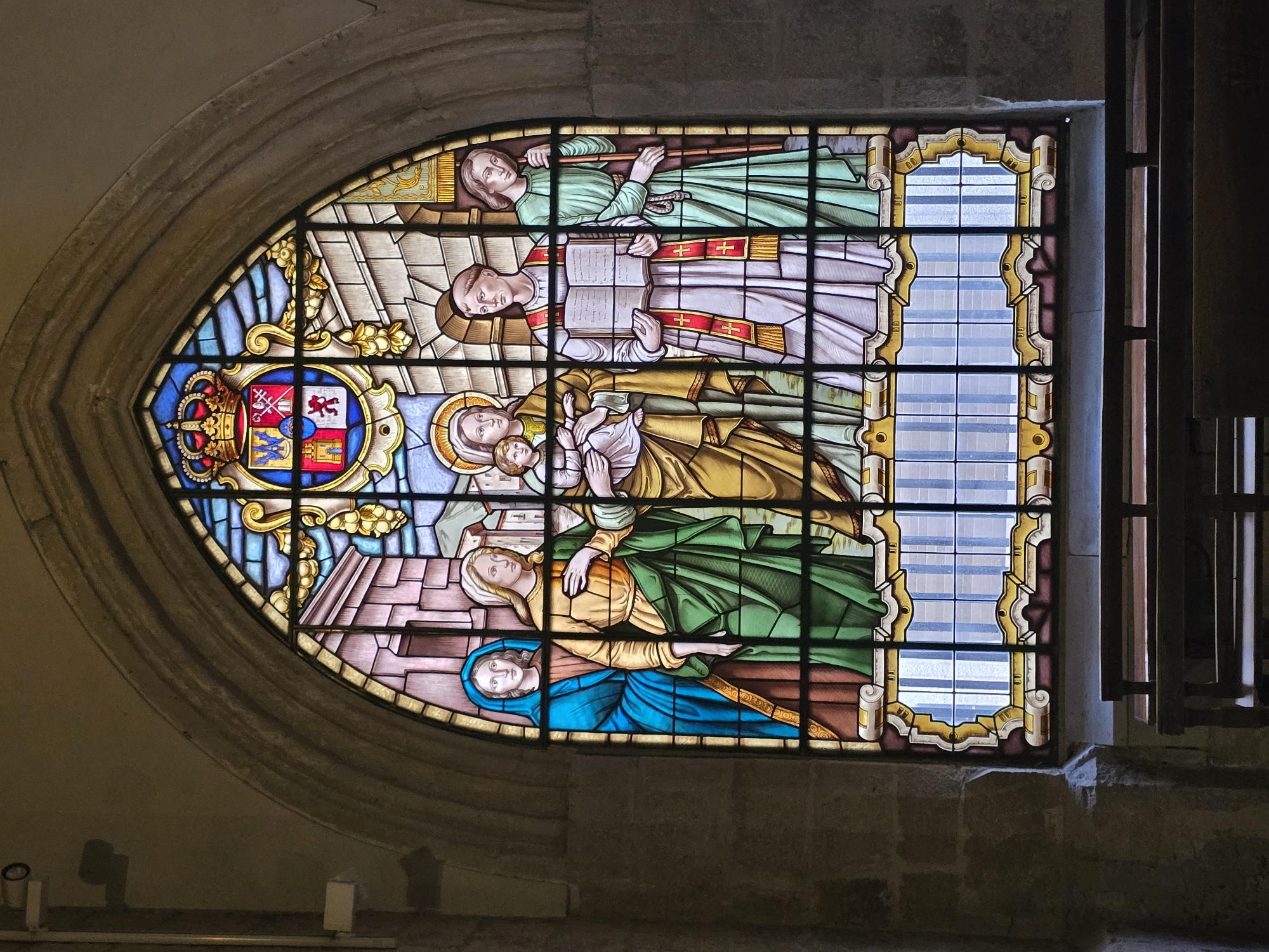 The image shows a stained-glass window depicting several figures in religious attire, located in the Cathedral of Jaén, Spain. The window is framed by stone architecture. In the foreground, a wooden bench is visible. The stained glass depicts a coat of arms at the top, with several figures below. From left to right, there is a woman in a blue robe holding a baby, a woman in a gold robe with a green sash, a figure in a white robe with a red and gold stole holding an open book, and a figure in a green robe holding a staff. The figures are rendered in a traditional artistic style with vibrant colors.