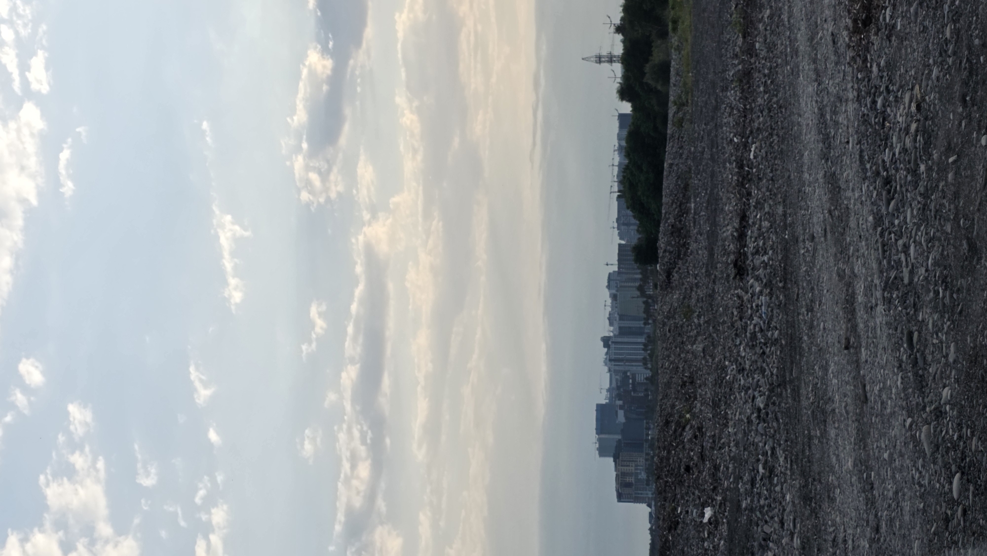 This image captures a broad, horizontal view of the outskirts of Batumi, Georgia, featuring a stark contrast between a desolate foreground and a developing urban landscape in the distance. The foreground is dominated by a vast expanse of dark, grey gravel and scattered larger stones, suggesting an undeveloped or industrial area, possibly a construction site or a landfill. A few white pieces of debris are visible among the stones on the left side of the frame.
In the middle ground, a dense line of green trees forms a natural barrier, separating the barren foreground from the distant city. Behind the trees, a cluster of modern, multi-story buildings, indicative of urban development, rises against a soft, cloudy sky. These structures vary in height and design, suggesting a mix of residential and commercial properties. Construction cranes are visible among the buildings, signifying ongoing development. To the far right of the mid-ground, a tall electrical pylon stands prominently against the sky, hinting at the city's infrastructure.
The sky above is a pale blue, adorned with scattered, wispy white clouds that are softly illuminated, suggesting it might be late afternoon or early morning. The overall impression is one of a city undergoing significant growth, with the raw, undeveloped land in the foreground highlighting the transition from a less developed area to a burgeoning urban center. The scene appears devoid of any people or animals.