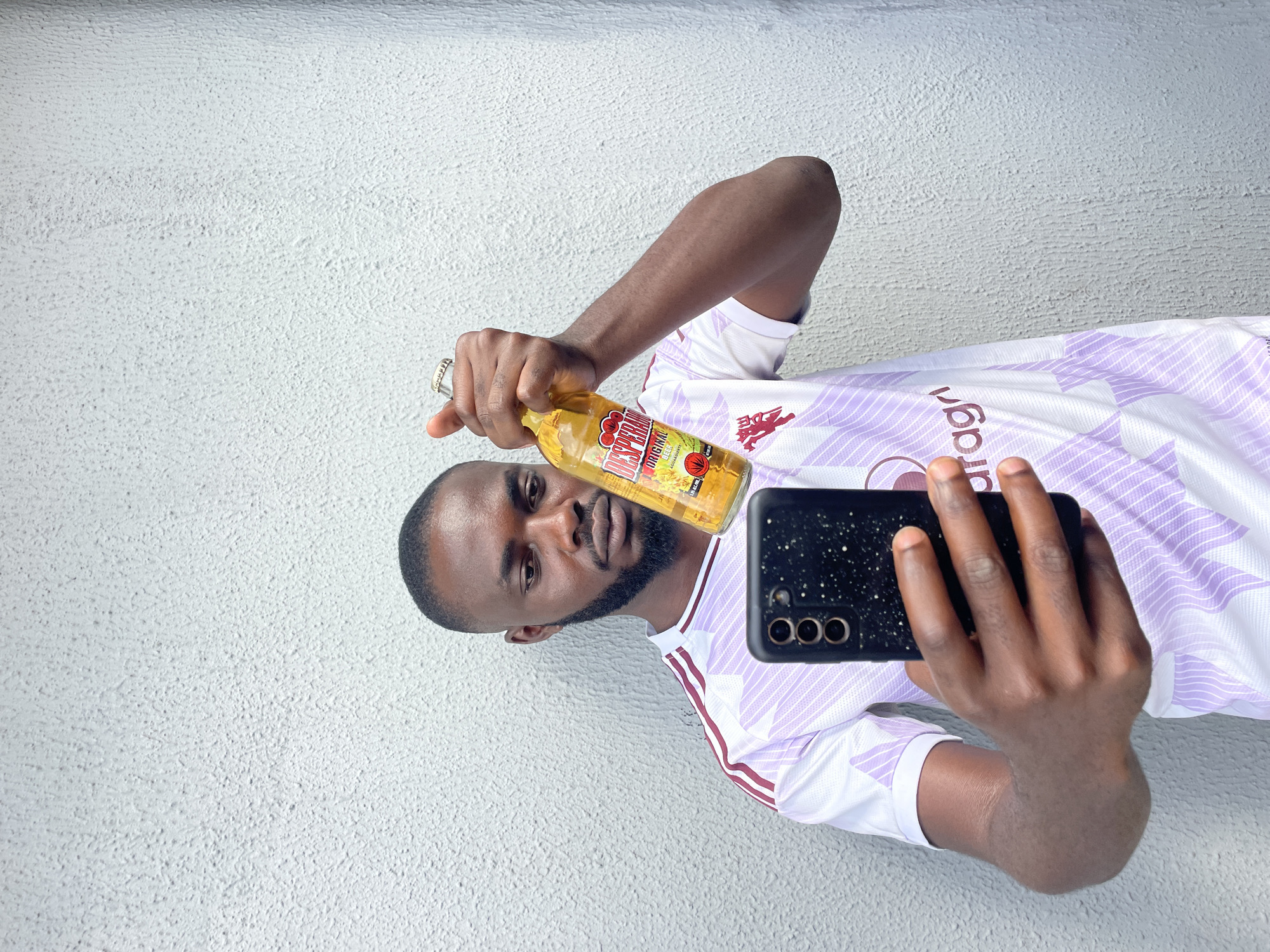 A man is taking a selfie outdoors, holding a bottle of Desperados Original Beer in his left hand and a black smartphone in his right. He is wearing a white and purple striped jersey. The background is a textured light-colored wall. The man appears to be in good spirits, with a slight smile. The lighting suggests it is daytime. The text "DESPERADOS ORIGINAL BEER" is visible on the bottle. The location is identified as Jos, Nigeria.