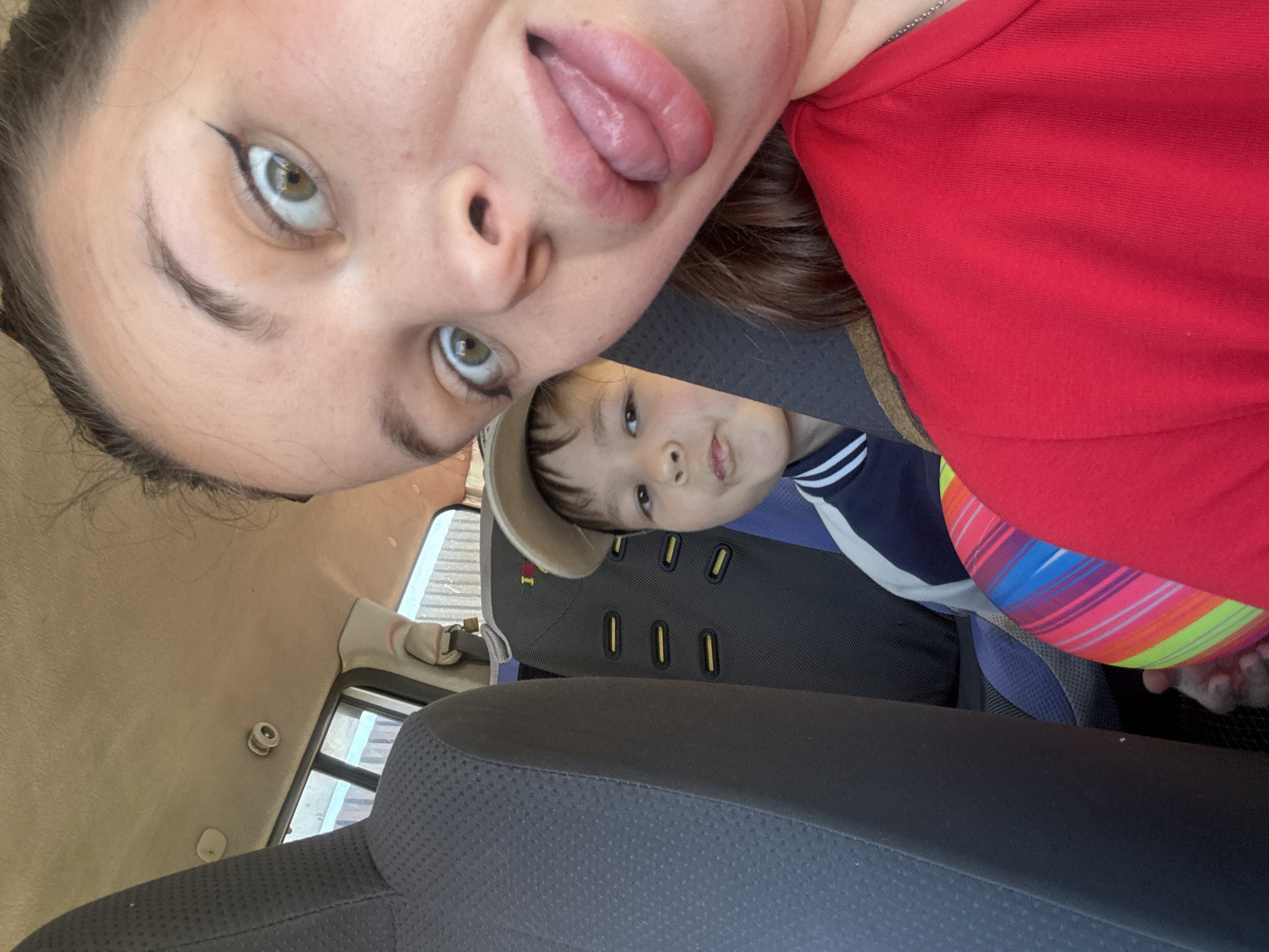 This media file captures a selfie of two individuals inside a vehicle. The primary subject is a young person with green eyes and dark hair, wearing a red shirt and makeup, sticking their tongue out. They are positioned in the foreground. Behind them, a young child wearing a light-colored hat and a dark blue and white striped shirt is visible, looking towards the camera. The child is seated in a car seat.
The setting appears to be the interior of a car, with car seats and the vehicle's headliner visible. Outside the car windows, a blurry glimpse of a somewhat overcast or diffused daylight can be observed. The overall tone of the image is casual and playful, suggested by the subjects' expressions and the selfie format.
There is no visible text in the image. The location context provided is Bely Yar, Russia. The time of day and weather conditions are not definitively discernible from the visual cues.