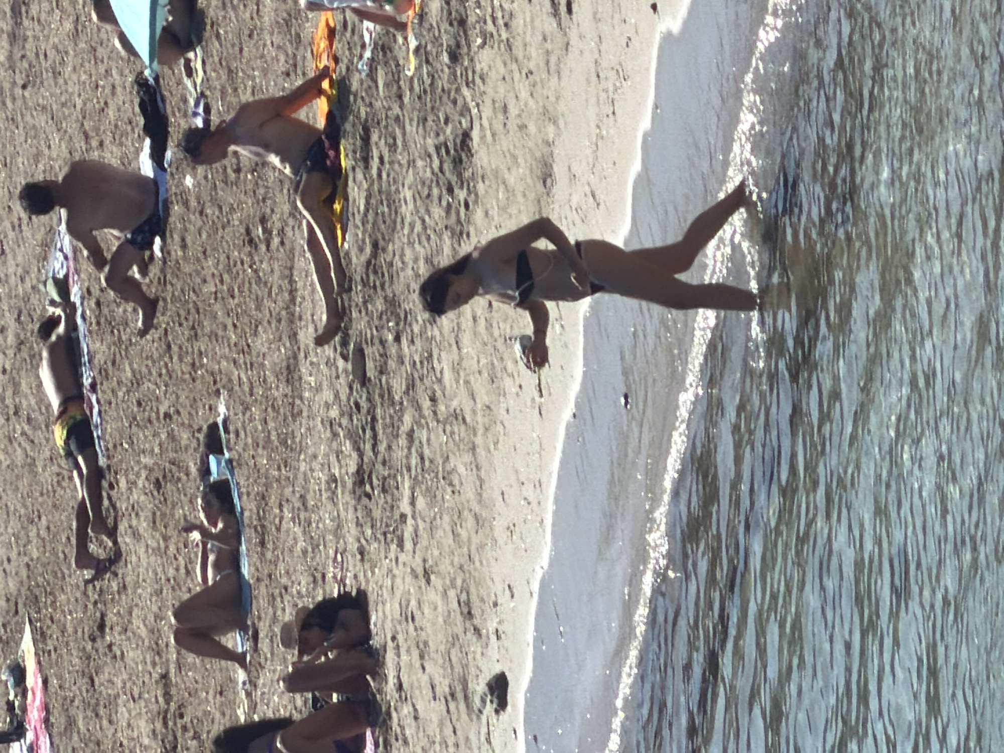 The image shows a group of people on a beach in Eivissa, Spain. The scene is captured from a high angle, with the ocean on the right and the sandy beach on the left. Several individuals are visible, most of whom are wearing swimwear.
On the left side of the image, several people are positioned on the sand. One person, wearing dark shorts, is lying down with their head towards the ocean. Another person, wearing colorful shorts, is standing and facing away from the camera, appearing to be walking towards the water. A third person, also in dark shorts, is partially visible further up the beach.
In the center of the image, a person is depicted in a swimming pose, with their body partially submerged in the water. Their face is not clearly visible.
The overall atmosphere appears to be relaxed, suggesting a day at the beach. The lighting indicates it is daytime, likely sunny given the bright reflections on the water. There is no visible text in the image.