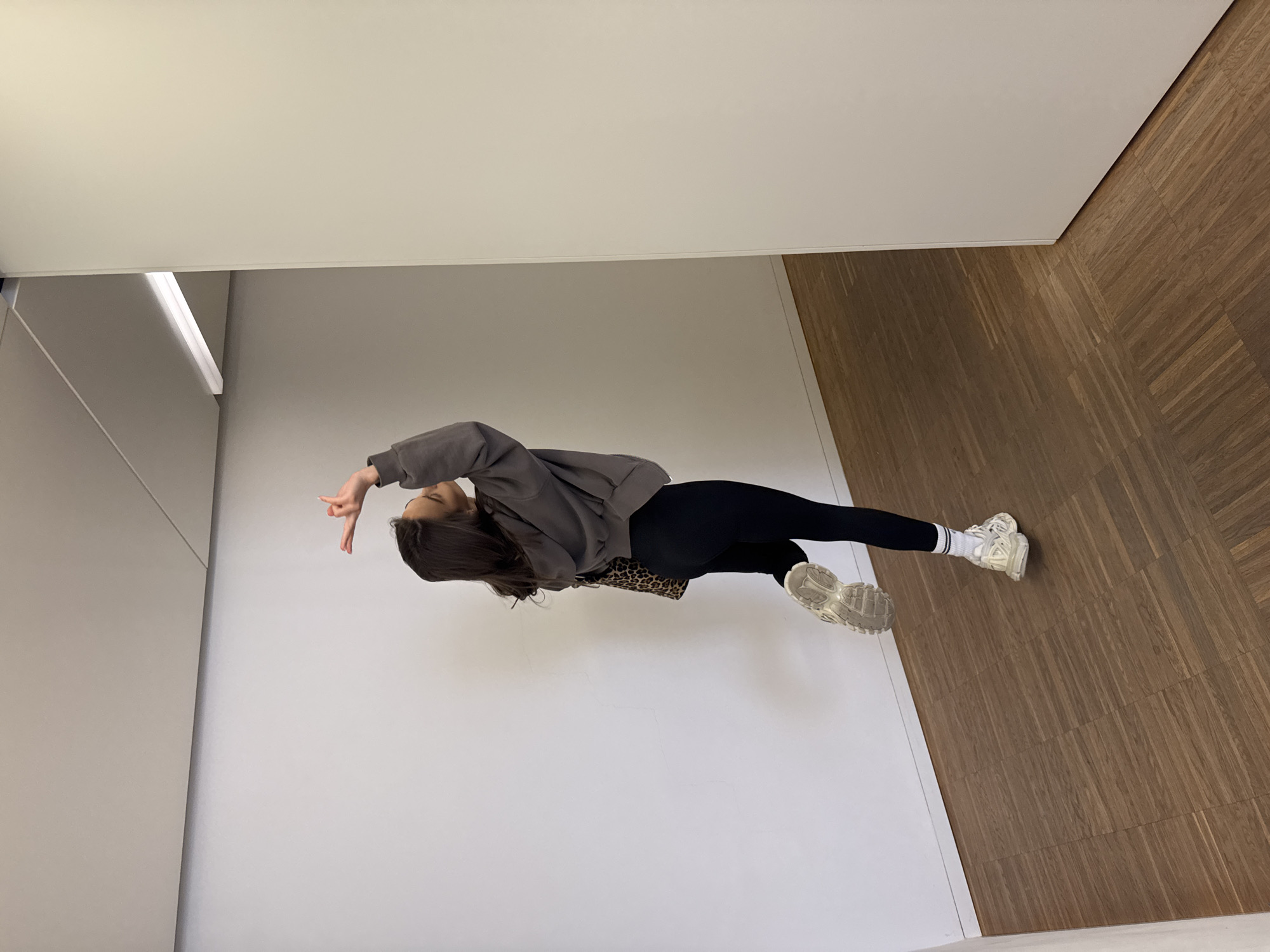 A young woman stands in a brightly lit, modern interior with white walls and a wooden parquet floor. She is wearing a gray sweatshirt, black leggings, and white sneakers with white socks. She has long, dark hair and is partially turned toward the camera, raising her right hand to make a hand gesture. A leopard-print purse is slung over her shoulder. The room appears to be a hallway or foyer, with a blank white wall and a corner visible in the background. The city and specific location are unknown.