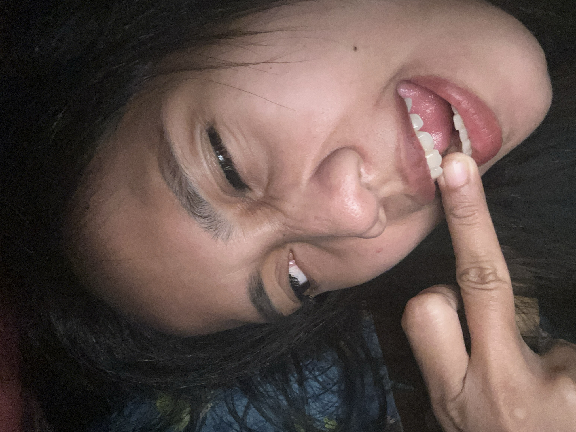 The image is a close-up, low-light, selfie-style photograph of a person's face and hand. The subject is a young female with dark hair and medium-toned skin. She is looking towards the camera with a playful or mischievous expression, her left eye is squinting, and her tongue is slightly protruding, touching her teeth. Her right index finger is in her mouth, touching her teeth. The background is dark and appears to be indoors, with some blurry blue and dark colors suggesting a patterned fabric or background. There is no indication of the city of Depok, Indonesia, in the image.