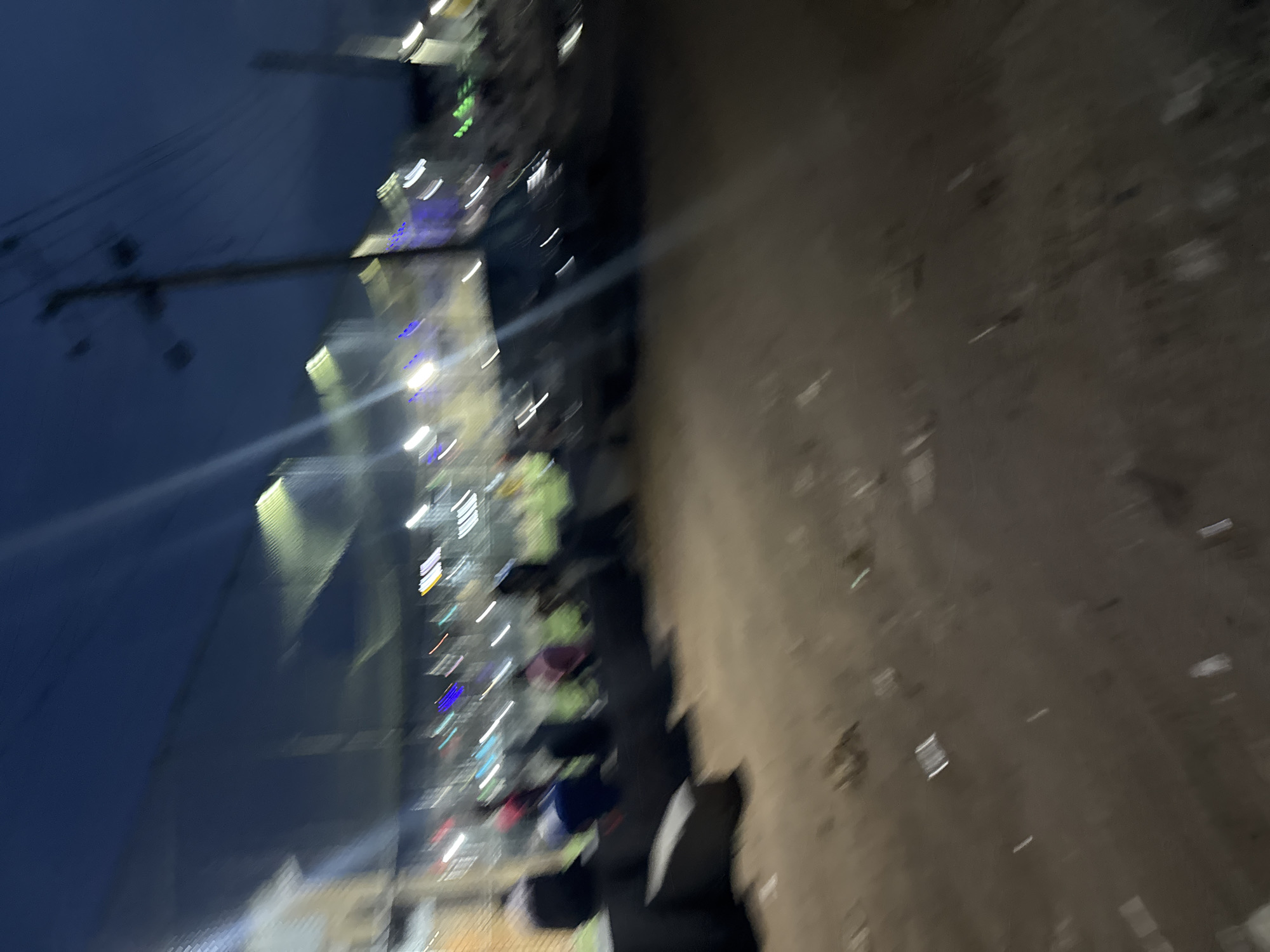 The image captures a very blurry, low-light scene at night or late evening, oriented 90 degrees clockwise. When viewed correctly, the upper portion shows a dark blue night sky with a prominent utility pole and overhead wires.
Below the sky, a row of illuminated structures or buildings stretches horizontally across the middle ground. These structures emit various artificial lights, primarily white, blue, and some green, which appear as long, streaky blurs due to motion or focus issues. The vibrant light streaks suggest an active area.
In front of these illuminated structures and extending towards the foreground, indistinct, blurred shapes are visible. These shapes appear to be people or objects, with hints of different colors like light green, blue, and dark tones, suggesting individuals gathered or engaging in an activity.
The foreground and right side of the image consist of an unpaved, light brown ground, likely dirt or earth, with scattered pieces of white and dark debris or litter.
The overall impression is that of a bustling outdoor setting, possibly a street or informal market area in Ihiagwa, Nigeria, characterized by artificial lighting and human presence, but captured with significant motion blur, rendering most details indistinct. No visible text is present.
