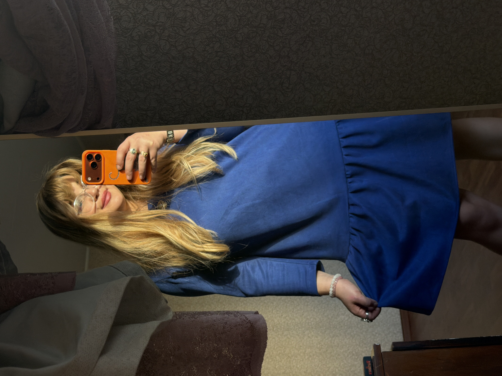 A blonde woman with glasses takes a mirror selfie indoors. She holds an orange smartphone with three camera lenses in her right hand. She wears a blue dress with long sleeves and a ruffled hem. She is wearing multiple rings on her left hand and a beaded bracelet on her left wrist. The background includes a textured dark wall, a draped cloth, and a wooden cabinet. The location is likely an apartment or home interior. There is no definitive evidence indicating the city of Naberezhnye Chelny, Russia.