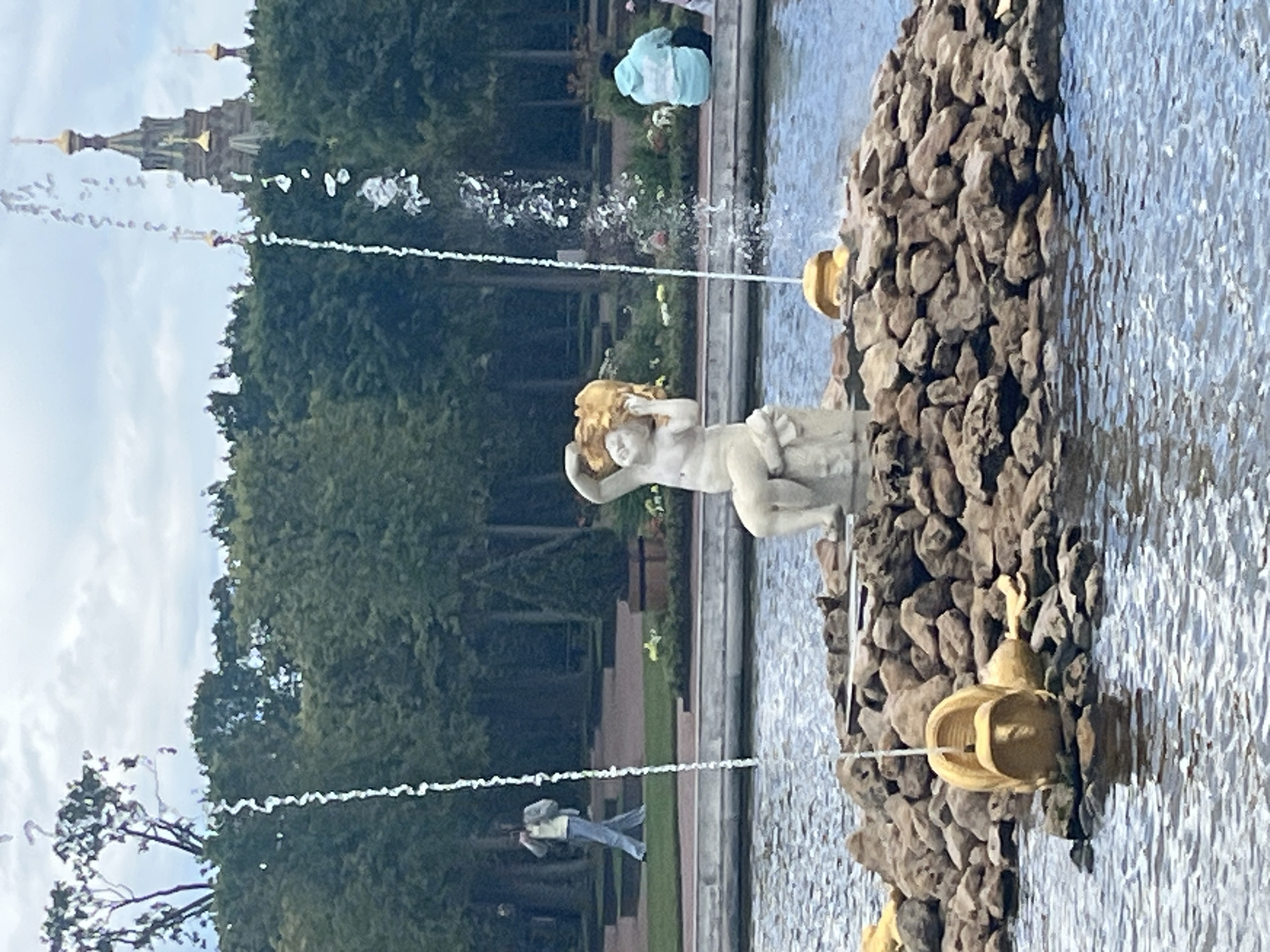 The image depicts a fountain in Peterhof, Russia, with a central sculpture of a cherub holding a golden object. The cherub is seated on a rocky mound adorned with golden accents, and water streams down from above. Several other golden elements, resembling bags or satchels, are scattered on the rocks around the mound. The fountain is set within a body of water, with lush green trees forming a backdrop. In the distance, a building with golden domes is visible. Two individuals are present: one person in the mid-ground, walking on a path and holding a phone, and another person in the background, wearing a light blue hoodie and sitting. The sky is overcast.