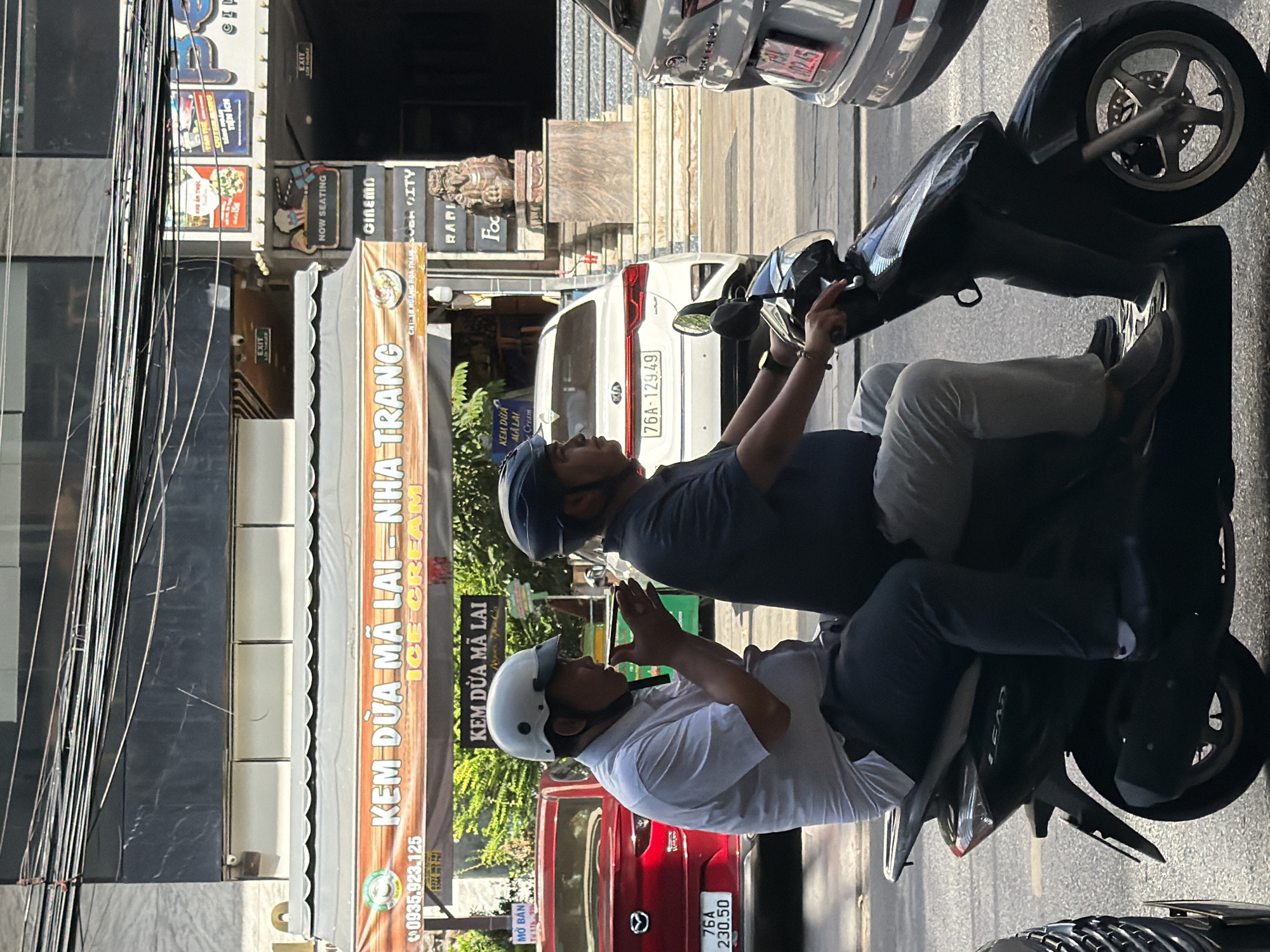 This is a street-level, eye-level, medium shot image captured in Nha Trang, Vietnam, likely on a sunny day given the strong shadows. The primary focus is on two men riding a black scooter. The man in front, wearing a white helmet and a light-colored short-sleeved shirt, is holding a small, dark object in his hands near his face, possibly a phone or a small item. He is looking slightly to his left. The man behind him, wearing a dark helmet and a dark short-sleeved shirt, is looking forward. He is wearing a watch on his left wrist.
The scooter has a license plate with the numbers "LEAD" visible on the rear. Behind the scooter, traffic is visible. A red Mazda CX-5 SUV is parked or stopped, with a license plate reading "76A 230.50". Further back, a white Kia sedan is also present, with a license plate "76A 129.49". To the right, the rear of a grey SUV with a license plate "102.45" is visible.
Above the street level, a building with multiple floors is seen. On the ground floor, there is a sign for "KEM DUA MA LAI - NHA TRANG ICE CREAM" with Vietnamese text and a phone number. Another sign next to it indicates "CINEMA". Above the cinema entrance, there are posters and signs related to a movie theater, including a "NOW SEATING" sign. Electrical wires crisscross above the street. The scene suggests a bustling commercial area in Nha Trang.