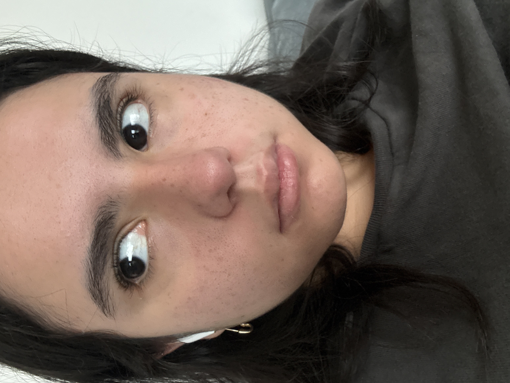 This is a close-up portrait of a person's face, rotated 90 degrees counter-clockwise, suggesting the subject is lying down. The person has fair skin with numerous visible freckles across the nose and cheeks. They have dark, wide-open eyes with visible reflections in the pupils, gazing directly into the camera. Their eyebrows are dark and well-defined. The lips are naturally pinkish, slightly parted, and appear full. Dark hair frames the face and spills onto a dark gray, textured fabric, which appears to be clothing or bedding. A small, gold-colored earring is visible on the earlobe on the right side of the face (left side of the image). A white object, possibly an earbud, is partially visible near the left ear (right side of the image). The lighting is soft and even, typical of an indoor setting. No visible text is present. The expression is neutral, conveying a sense of direct engagement or contemplation.