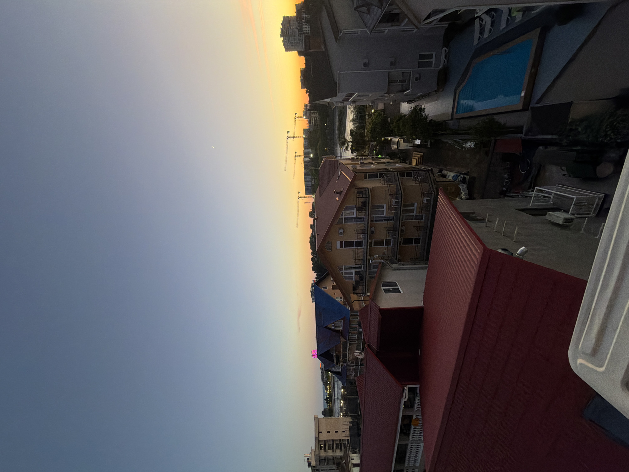 This is a high-angle, full-shot photograph of a coastal town at sunset, likely in Sirius, Russia. The sky is a gradient of soft blues and oranges, with a sliver of a moon visible. In the foreground, several buildings with red and dark-colored roofs are prominent. One building, painted yellow, has multiple balconies with white railings and air conditioning units. To the right of this building, a grey apartment building stands, and below it, a bright blue swimming pool with lounge chairs is situated. Further back, a body of water is visible, beyond which are more buildings and construction cranes dotting the horizon. The overall atmosphere is serene, with the fading light casting a warm glow over the scene. There are no people or animals visible in the image.