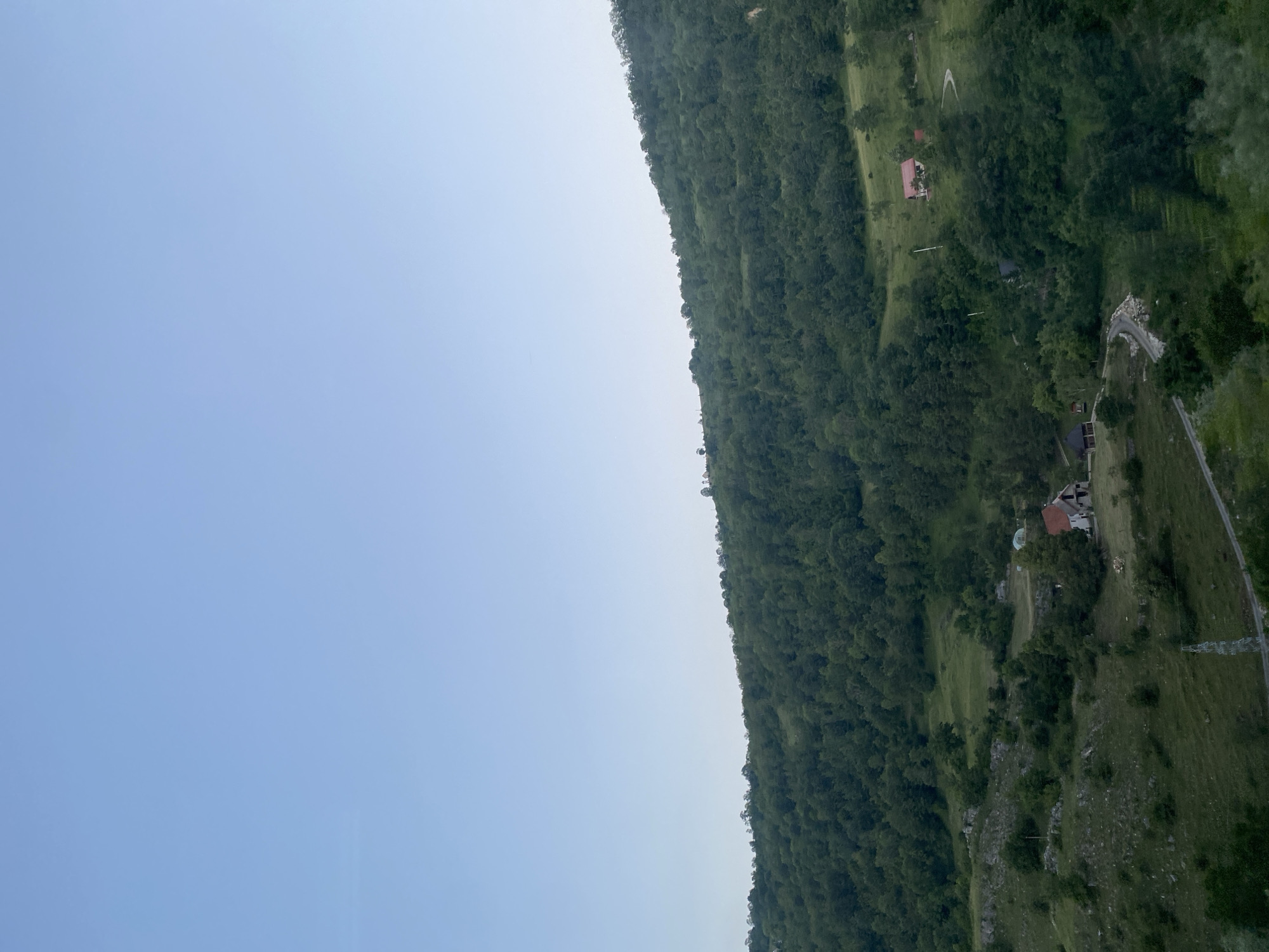 A wide shot captures a serene, hilly landscape under a clear, pale blue sky. The terrain is covered with lush green trees and vegetation, with a few scattered houses nestled within the rolling hills. The perspective is from a slightly elevated position, looking down and across the verdant slopes. The image appears to be taken in the vicinity of Šavnik, Montenegro, a region known for its natural beauty and mountainous terrain. No people or animals are visible in the frame. The scene is peaceful and untouched by human activity, showcasing the raw beauty of the natural environment.