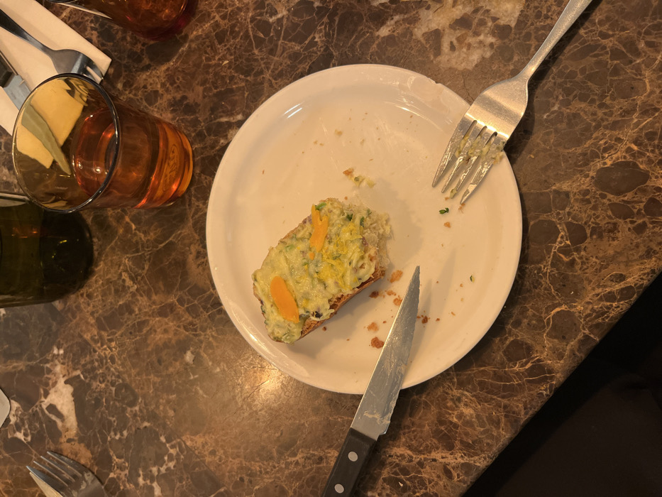 An overhead shot captures a nearly finished meal on a marble surface. A white plate holds a partially eaten slice of toast topped with a yellow-green spread, possibly an egg salad. A stainless steel fork and knife lie on the plate, alongside the toast. Two drinking glasses, one with an amber liquid and another with a dark green liquid, are present. Silverware sits near the corner of the table, completing the restaurant setting.