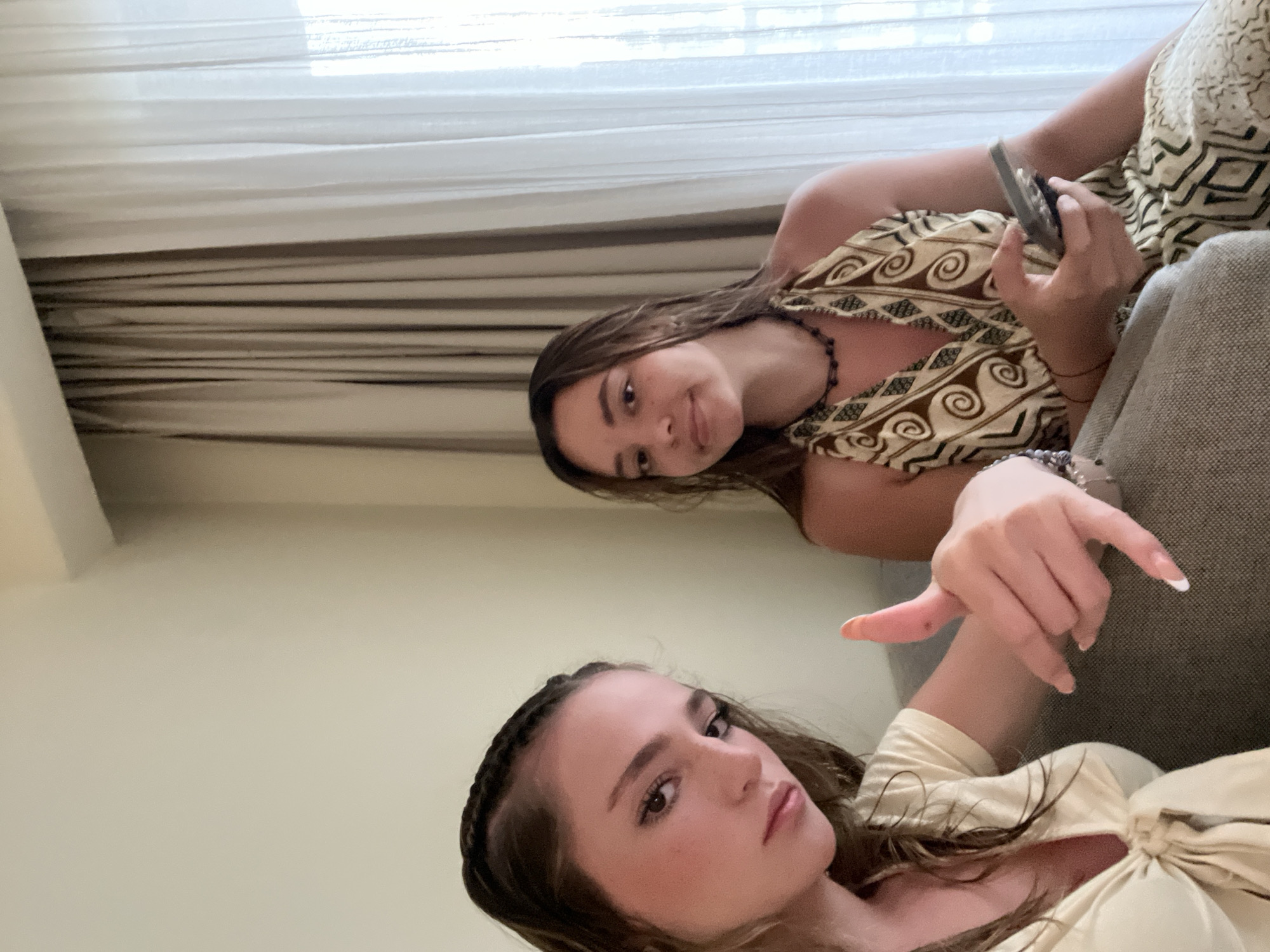 Two young women pose for a selfie indoors. The upper woman wears a brown and beige patterned top and holds a phone. The lower woman wears a pale yellow top and makes a hand gesture. They appear to be on a gray sofa. A curtain is visible in the background. The location is possibly Playa del Carmen, Mexico.