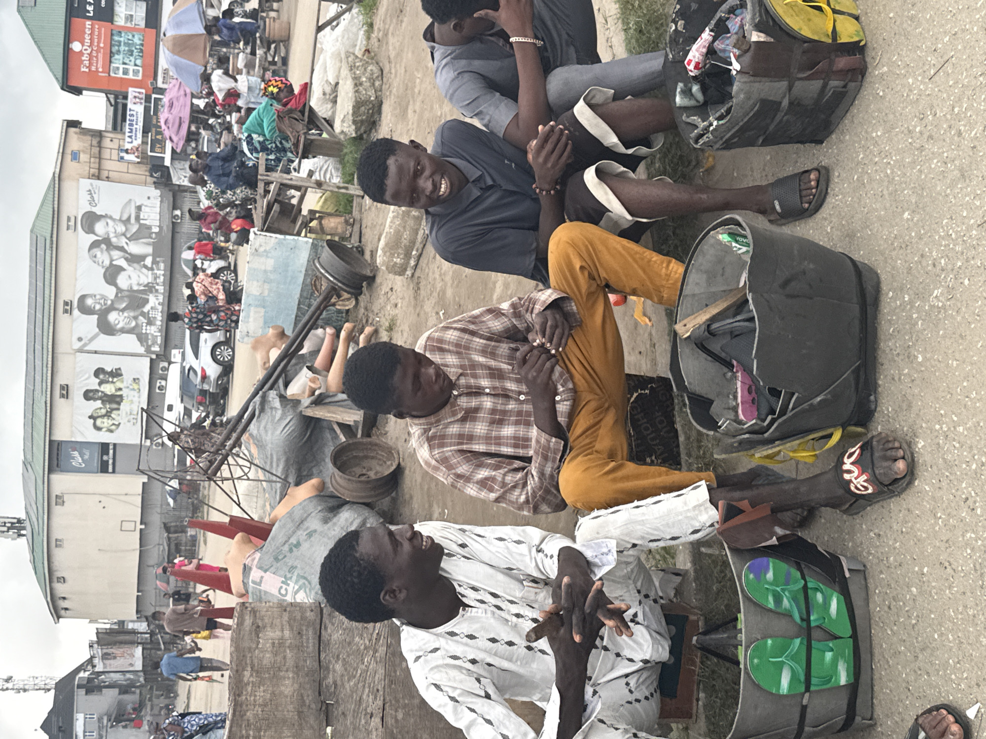 The image depicts a street scene in Satellite Town, Nigeria, under an overcast sky. Four young men are seated together on makeshift stools, appearing to be engaged in conversation or a transaction.
The man on the far left wears a white patterned traditional garment and is smiling. Next to him, a man in a plaid shirt and orange pants is looking down, holding something in his hands. The third man, in a dark blue polo shirt and patterned shorts, is also looking forward and appears to be speaking. The man on the far right, dressed in a grey shirt and pants, has his head bowed, with one hand resting on his knee.
In front of the men are several large, dark grey plastic buckets filled with various items. One bucket prominently displays a pair of green flip-flops with a purple pattern. Another bucket contains what appears to be clothing and other miscellaneous objects. To the right, another bucket is visible, also filled with items.
The background reveals a busy urban environment. A large building with "Class" and other text on a billboard is visible, along with signage for "FabQueen Hair & Couture" and "Lambest". Several vehicles, including cars and motorcycles, are present on the street, along with a variety of people walking and interacting. Umbrellas are scattered throughout the scene, suggesting it might be a market or a public gathering area. The ground is a mixture of dirt and pavement. The overall impression is one of daily life in a bustling Nigerian city.