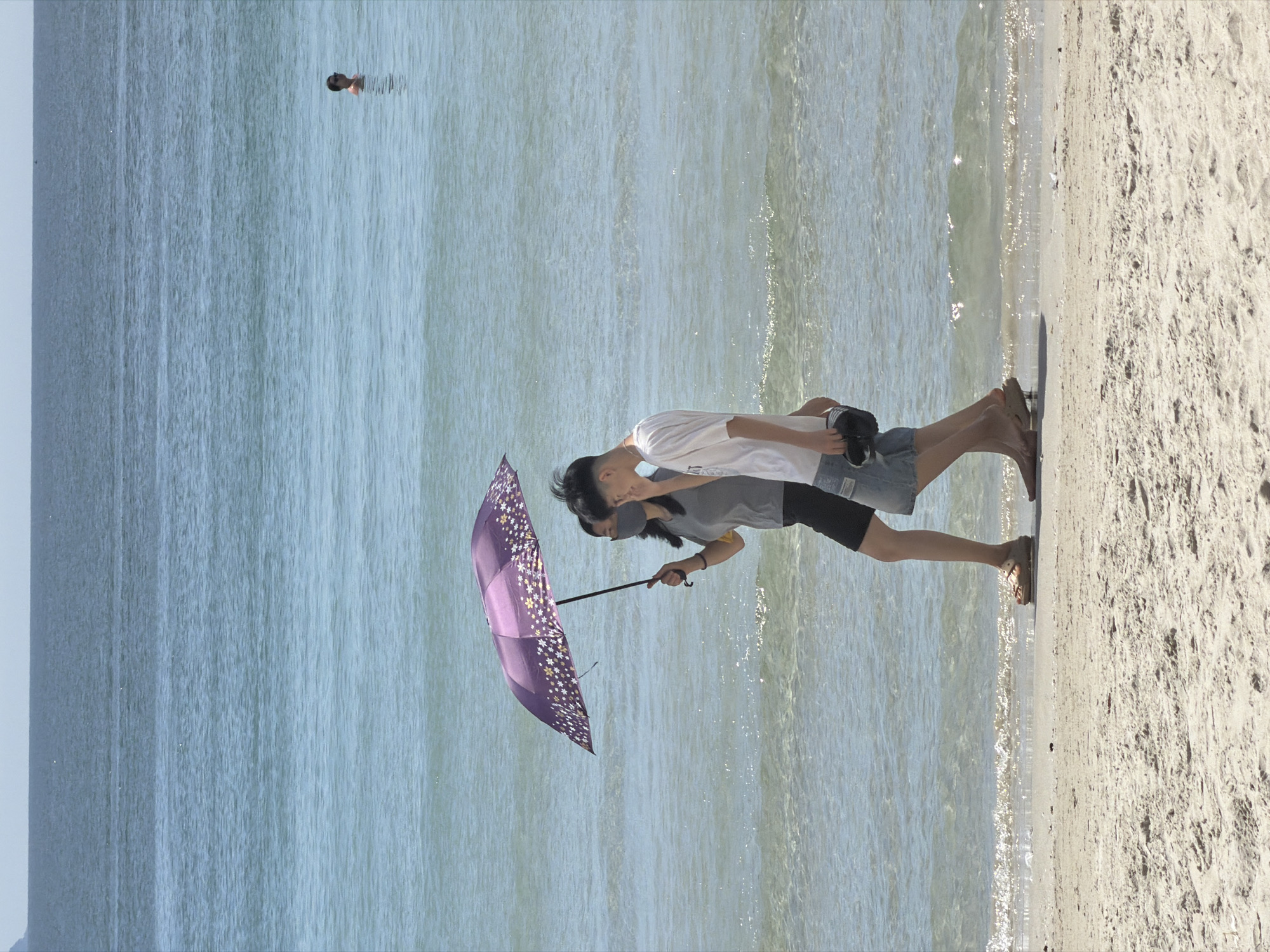 The image shows a sunny day at a beach in Dong Ninh Hoa Ward, Vietnam. Two individuals are walking along the shoreline, with one person holding a purple umbrella decorated with white flowers. The individual holding the umbrella is wearing a grey t-shirt and black shorts and is also wearing a face mask. The other individual, who appears to be younger, is wearing a white t-shirt and denim shorts and is carrying a pair of black shoes. The sea is calm and blue, with a person visible swimming in the distance. The sand is light in color and shows some texture from footprints. The sky is clear and light blue.