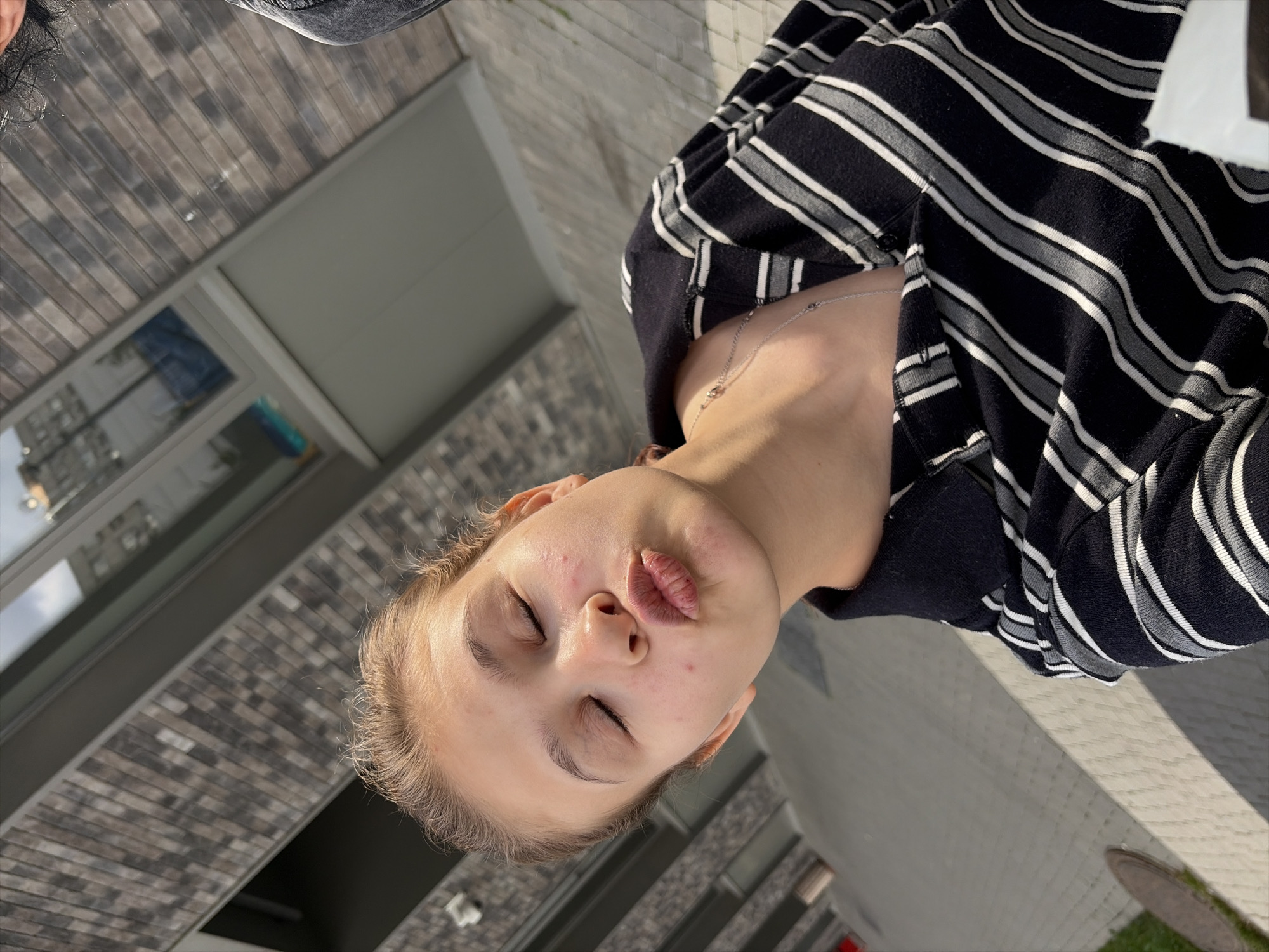 A young woman is taking a selfie outdoors. She is wearing a black and white striped shirt and a silver necklace. Her eyes are closed and her lips are pursed in a kiss. The background shows a modern building with brick siding and large windows. The ground appears to be paved with light-colored stones. There is a tree or bush visible on the left side of the image. The scene suggests a casual setting in an urban environment.