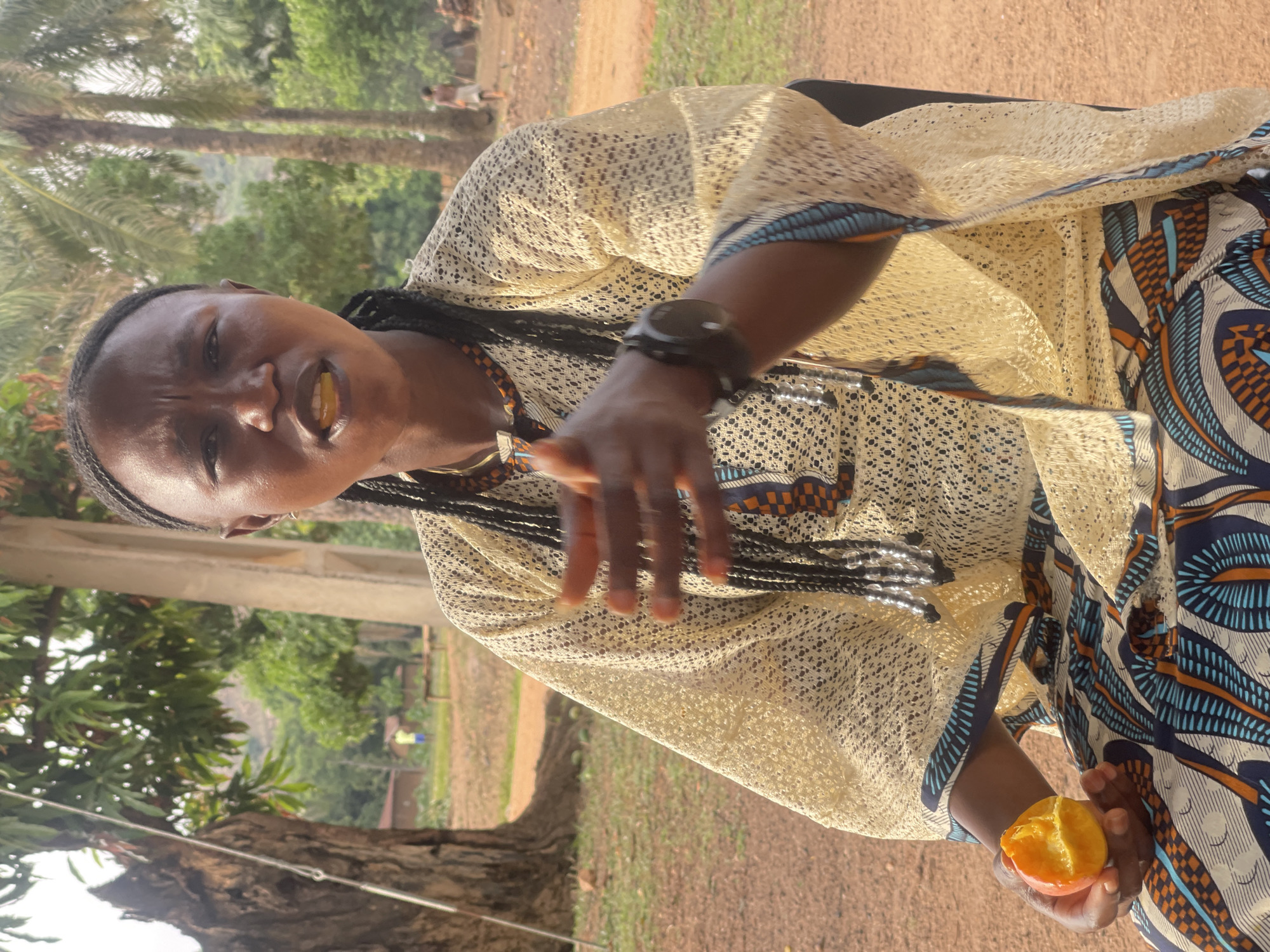 This image captures a close-up, outdoor portrait of a Black individual, likely female, set in Tabu, Nigeria. The subject has dark skin and her hair is styled in intricate cornrows. She is looking directly at the viewer with an intense, focused expression, her mouth slightly agape, revealing teeth and possibly a piece of fruit.
She wears a light, cream-colored, loosely woven or lacy overlay garment, paired with an undergarment featuring a vibrant African wax print pattern in blue, orange, and white. A dark watch is visible on her left wrist. Her left hand is outstretched in a gesturing motion, while her right hand holds a partially eaten, bright orange fruit, resembling a mandarin or a small orange.
The setting is a sunny, outdoor environment with a distinctive red-brown dirt ground. The background is lush with tropical foliage, including tall palm trees and other green trees, suggesting a rural or semi-rural landscape. Hints of distant structures or additional people can be faintly seen in the far background. The bright, direct sunlight indicates it is daytime, likely midday or early afternoon, under clear weather conditions. There is no visible text in the image.