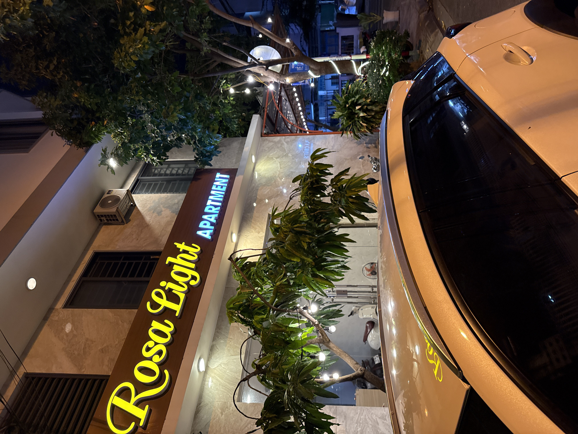 The image depicts the facade of a building at night, identified as "Rosa Light APARTMENT" in Nha Trang, Vietnam. The building features a prominent yellow illuminated sign displaying the name "Rosa Light" in a cursive font, followed by "APARTMENT" in blue neon lettering. The building's exterior is clad in marble-like tiles, and architectural details like windows with dark shutters and an air conditioning unit are visible.
In the foreground, the rear portion of a light-colored car is partially visible, with its roof reflecting the building's lights. The car appears to be parked on a street.
The environment includes several trees with lush green foliage, some adorned with string lights, creating a festive ambiance. The street lighting and the illuminated sign suggest it is nighttime. In the background, there are glimpses of other buildings, and street lights, indicating an urban setting. A white scooter is parked near the building. The overall scene conveys a welcoming and possibly commercial establishment in an urban environment at night.
