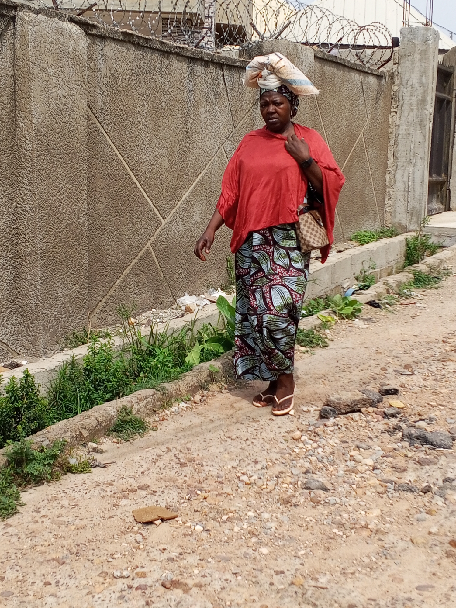 The scene depicts a woman walking on a dirt road in Adankolo, Nigeria. She is wearing a red top, a patterned skirt, and sandals, with a bag over her shoulder and a wrapped bundle on her head. The road is bordered by a concrete wall topped with barbed wire, and some vegetation grows along the base of the wall. The road surface is uneven and consists of dirt, gravel, and small stones.