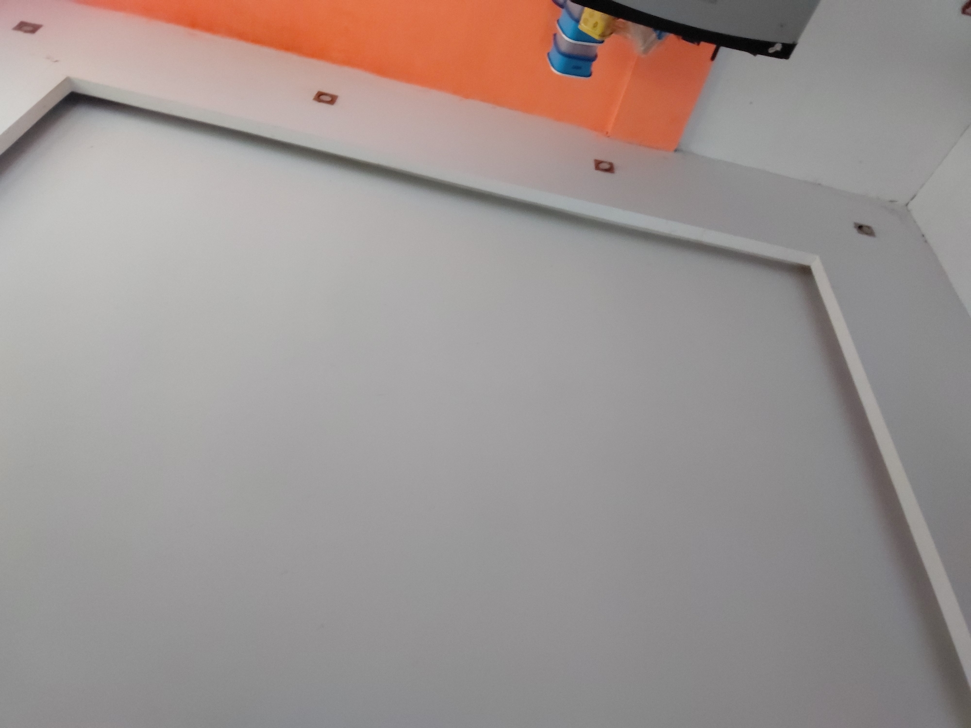 The image displays an upward-angled view of an interior room, focusing on the junction of walls and ceiling. The predominant subject is a large, unadorned light gray-white wall that occupies the lower and central portion of the frame. Above this main wall, a broad white architectural trim or border creates a recessed area, extending horizontally across the room.
Above this white trim, a striking section of wall or ceiling is painted a vibrant, solid orange color, providing a bold accent. Embedded within the white trim, and in the upper right corner of the image, are several small, square, dark brown or black objects. These appear to be either recessed lighting fixtures, electrical outlets, or mounting points.
In the upper center of the image, brightly colored items are suspended from what seems to be an overhead fixture. These include components in blue, yellow, and white, resembling clothing items, pegs, or parts of a children's mobile or toy. A dark, possibly black, rectangular object, which could be a television screen or its mounting hardware, is partially visible just above these hanging items. The upper right corner reveals another section of white wall or ceiling, showing some minor surface imperfections or hairline cracks.
The lighting appears to be soft and diffused, characteristic of an indoor setting with either artificial light or indirect natural light. There are no individuals present, and no explicit activity is occurring, although the hanging objects might suggest a domestic environment. No discernible text is visible anywhere in the image. The scene conveys a contemporary or minimalist aesthetic, punctuated by the vivid orange color. The location context is Santa Ana, Venezuela.
