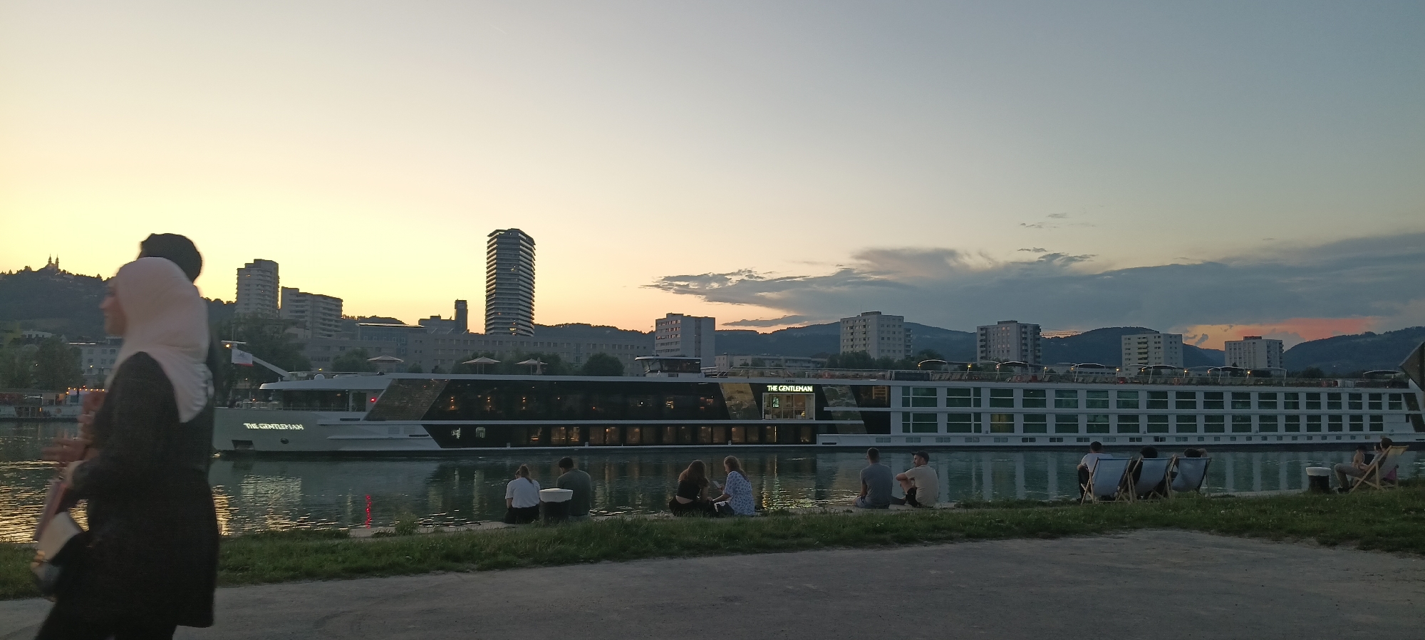 This is a wide-angle, eye-level photograph taken during sunset along the Danube River in Linz, Austria. The scene captures a picturesque view of the riverfront with a large cruise ship, identified as "THE GENTLEMAN," docked on the water. The ship features a modern, white design with numerous windows reflecting the ambient light.
In the foreground, a woman wearing a hijab and a dark outer garment is walking away from the camera, towards the left side of the frame. She is carrying a bag over her shoulder. Further along the riverbank, a group of people are seated on the grass, facing the water and the ship. These individuals appear to be enjoying the evening scenery. Some are sitting directly on the ground, while others are in lounge chairs.
The background displays the Linz cityscape, characterized by a cluster of buildings of varying heights, including a prominent, tall, modern skyscraper. The sky is a gradient of soft orange and yellow hues from the setting sun, transitioning to a pale blue and grey, with scattered clouds. The river itself reflects the sky and the lights of the city and the ship, creating a tranquil and inviting atmosphere. The overall impression is one of a serene evening by the river.