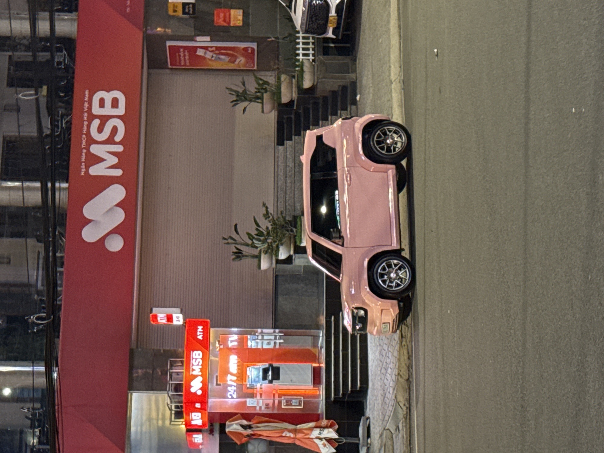 This nighttime image captures a street scene in Da Lat, Vietnam, characterized by artificial illumination. A striking pink compact car is prominently parked on the right side of a paved road, facing right. Its silver, multi-spoke wheels are visible. To the left of the car is a sidewalk, and beyond that, a multi-story building with a distinctive red facade.
The building appears to be a bank, identified by the large white letters "MSB" and the full name "Ngân Hàng TMCP Hàng Hải Việt Nam" (Vietnam Maritime Commercial Joint Stock Bank) displayed vertically on the red panel. Below this, at street level, is a glass-fronted section with bright signs indicating "ATM" and "24/7 ATM," confirming the presence of automated teller machines. Several dark steps lead up to an entrance of the building, flanked by potted plants. Another white car is partially visible, parked further up the street near some railing and more potted plants. Utility lines and cables are discernible on the far left. A folded orange and white umbrella or awning lies on the sidewalk near the ATM section. The street itself appears well-maintained, and the overall atmosphere is calm and illuminated by streetlights and building lights. No people are visible in the frame.