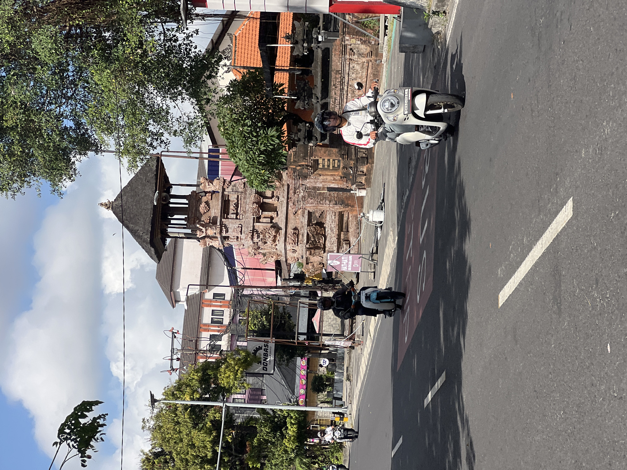 In Kuta, Indonesia, a bright, sunny day unfolds on a city street. Two individuals are riding scooters, one in the foreground and another further back. The person in the foreground is on a white scooter and is wearing a white and red jacket, dark pants, and a helmet. They are looking forward, focused on the road. Behind them, a second scooter rider is on a light blue and white scooter, wearing dark clothing and a helmet. This scooter is moving towards the background, away from the viewer.
The street is paved with asphalt and marked with white lines. A red lane marker, possibly for bicycles or a dedicated lane, is visible on the road. Lush green trees line the sides of the street, their leaves casting shadows on the asphalt.
Dominating the background is a traditional Balinese temple gate, constructed from reddish-brown stone. It features intricate carvings and a thatched roof. Behind the temple, buildings with orange tiled roofs and pink and white facades are visible, suggesting a commercial or residential area. A sign for "DONBASE CAR CARE SERVICE" is also present, indicating businesses in the vicinity. The sky is a vibrant blue, dotted with fluffy white clouds. The overall scene depicts a typical day in a bustling Indonesian city, with a blend of traditional architecture and modern life.
