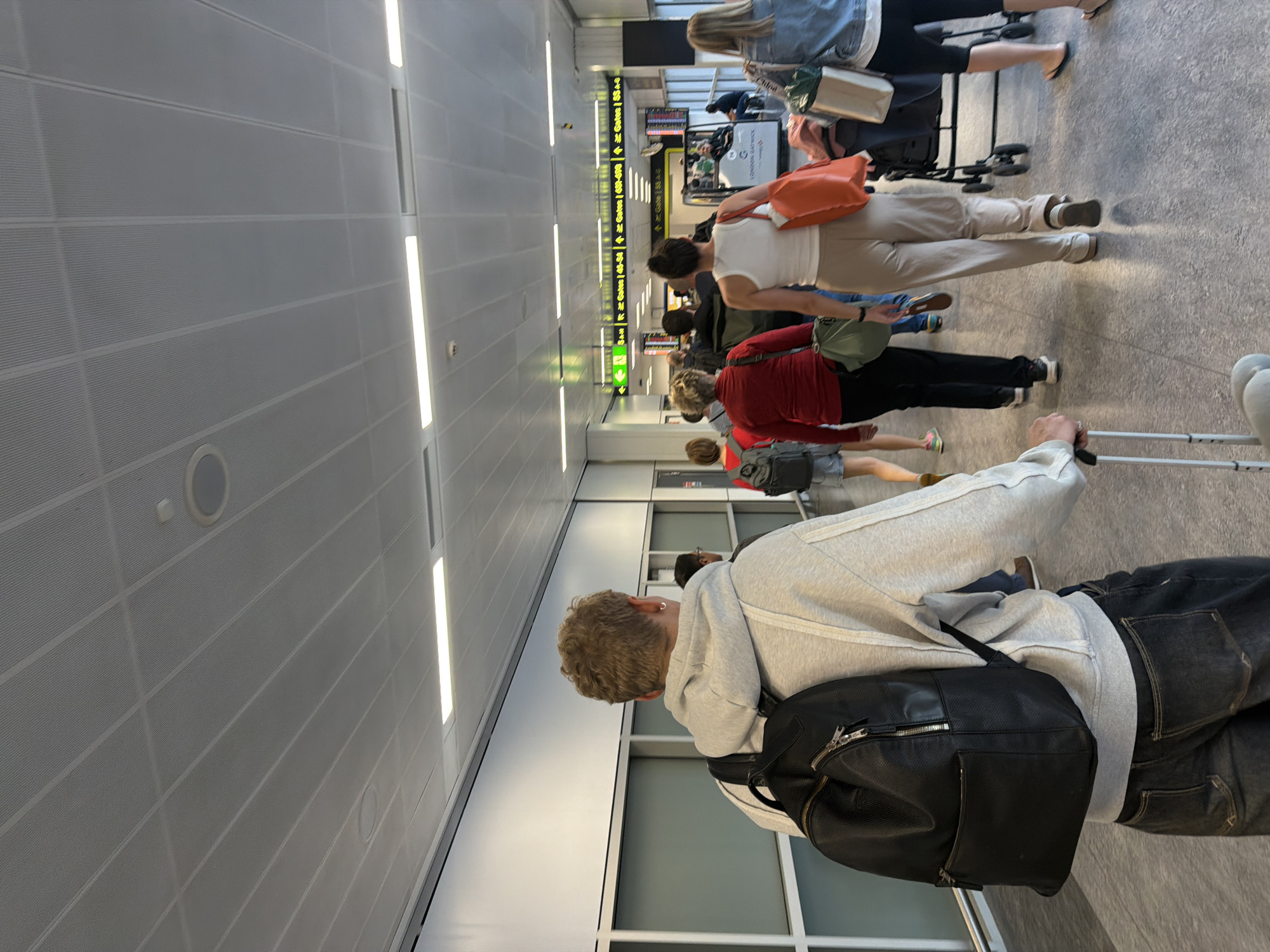 The image depicts a view from behind a person wearing a grey hoodie and black backpack, walking down a brightly lit corridor in an airport. Several other people are also walking in the same direction, some with luggage. Overhead, fluorescent lights illuminate the metallic ceiling panels. Green signs with white text indicating gate numbers and directions are visible in the distance. The floor is a speckled grey tile. The location is the transit area of London Gatwick Airport, Crawley, United Kingdom.