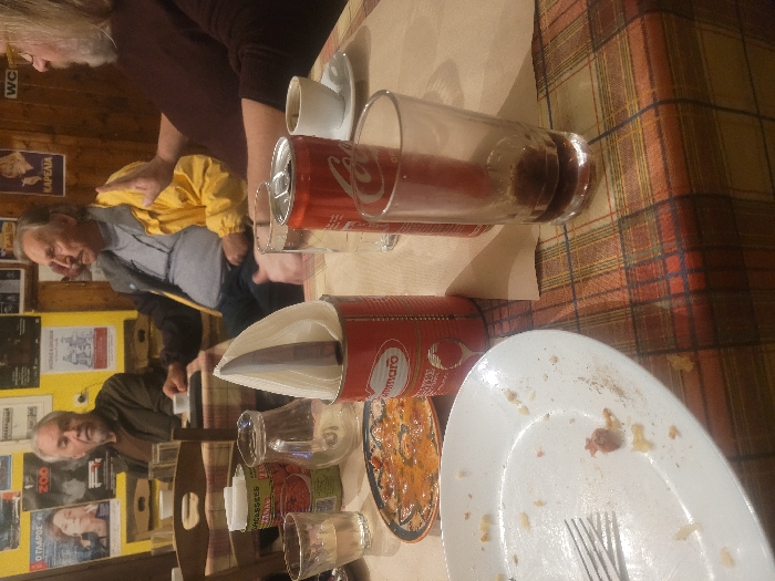 The image depicts three older adult males seated around a plaid-covered table in a casual eatery or tavern in Κωστήτσι, Greece. In the immediate foreground, a white ceramic plate with food residue and a silver fork is positioned next to a clear glass with a small amount of liquid. A decorative ceramic plate containing an orange-red dish is visible behind the white plate. A red San Marzano branded tomato can, repurposed as a napkin holder, stands upright on the table, containing white paper napkins and a dark, cylindrical object. To the left of the napkin holder, a clear glass pitcher is present. On the right side of the table, a red Coca-Cola can, an empty clear drinking glass, and a white coffee cup with a dark liquid residue on a saucer are visible.
The man on the far left has grey hair and a beard, wearing a brown jacket, and is looking towards the right side of the frame. The man in the middle has a mustache and is wearing a yellow jacket over a darker garment, also looking right. The man on the far right, partially visible, has long grey hair and a beard, wearing a dark reddish-brown long-sleeved shirt, with his right hand raised in an open-palm gesture, suggesting active communication. The background features light yellow walls partially covered with dark wood paneling, adorned with various posters and signs, including text such as "Ο ΓΑΡΟΣ", "ZOO", and "WC".