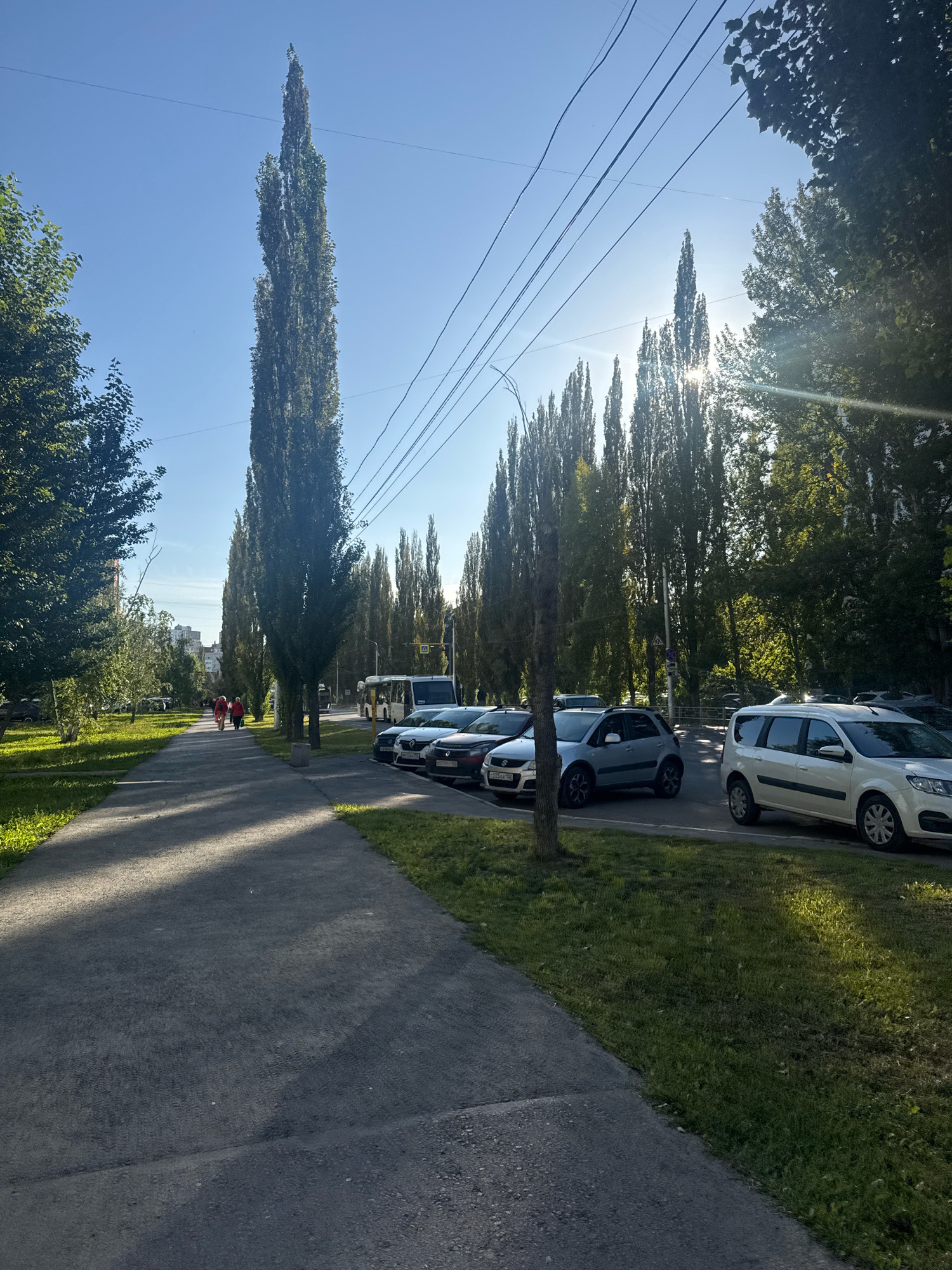 This image shows a street scene in Ufa, Russia. A paved sidewalk runs alongside a road lined with tall, narrow trees. On the sidewalk, two people in red jackets are walking away from the camera. Parked cars line the side of the road, and a bus is visible in the distance. The sun is shining brightly, casting long shadows across the sidewalk and illuminating the trees. The sky is clear and blue.