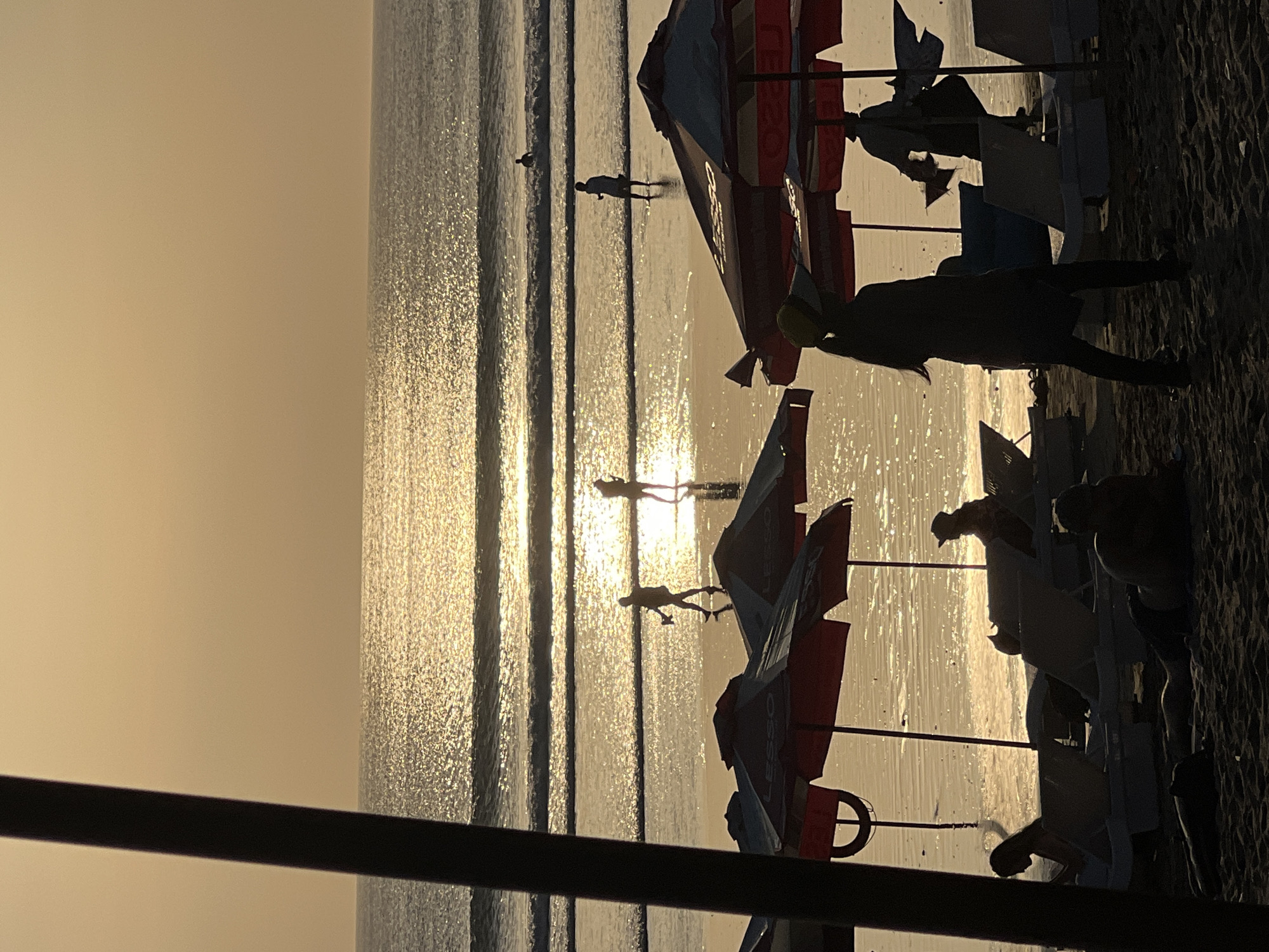 The image depicts a beach scene at sunset in Kuta, Indonesia. The sky is a soft orange hue, with the sun's reflection creating a shimmering path across the ocean waves. Several people are visible on the wet sand, walking and standing near the water's edge, silhouetted against the bright reflection. In the foreground, beach umbrellas and lounge chairs are set up on the sand. Some individuals are seated on the chairs, while others are walking in the sand or wading in the shallow water. The overall atmosphere is serene, with the golden light of sunset casting long shadows.