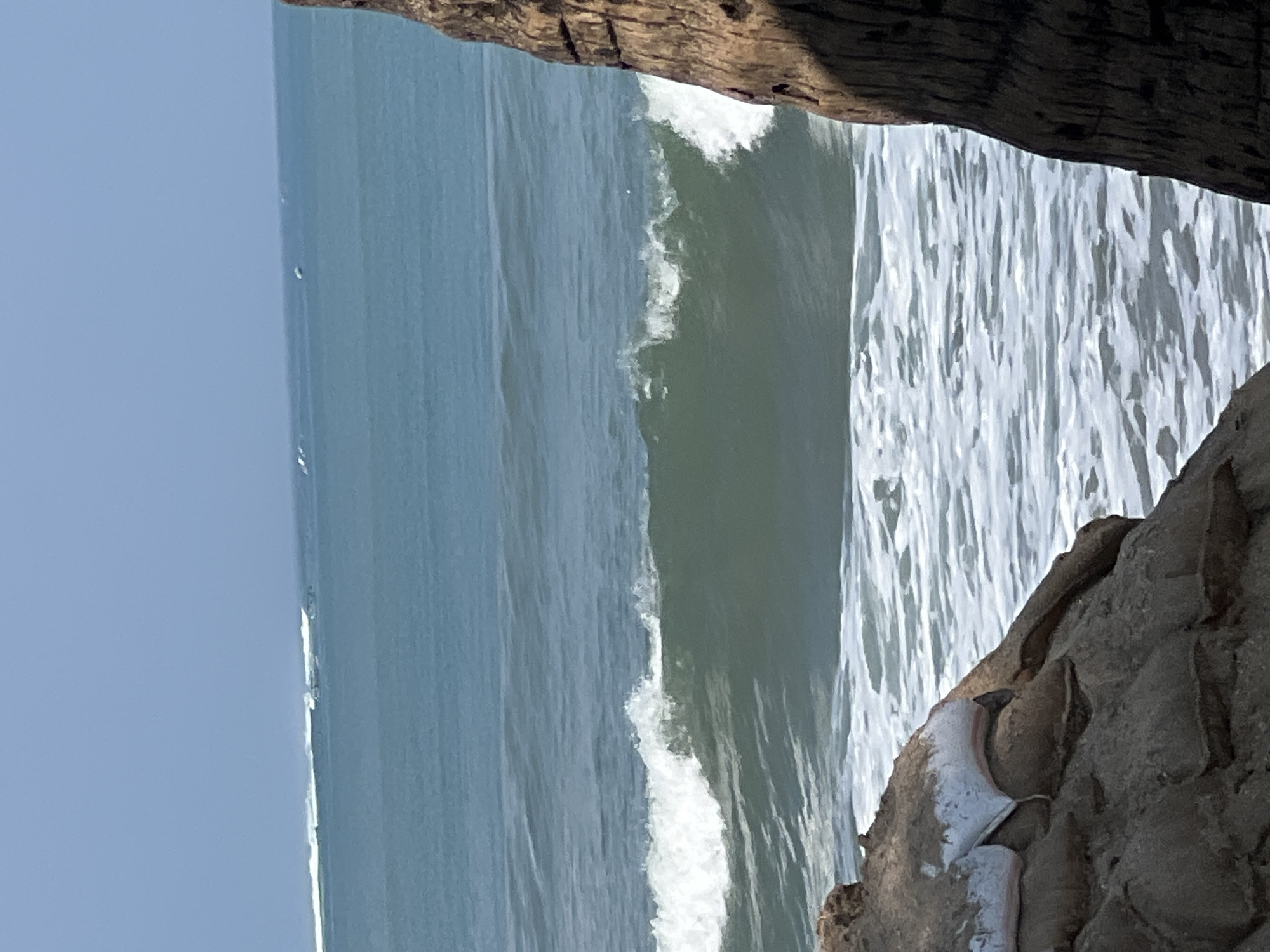 This is an eye-level, medium shot of waves breaking on the coast of Kuta, Indonesia. The ocean is calm with gentle swells in the distance and a larger, breaking wave in the foreground, creating white foam. The foreground is framed by two large, rough, dark brown rock formations. In the bottom left corner, a pile of sandbags is visible, covered in what appears to be white tarpaulin. The sky is clear and blue.