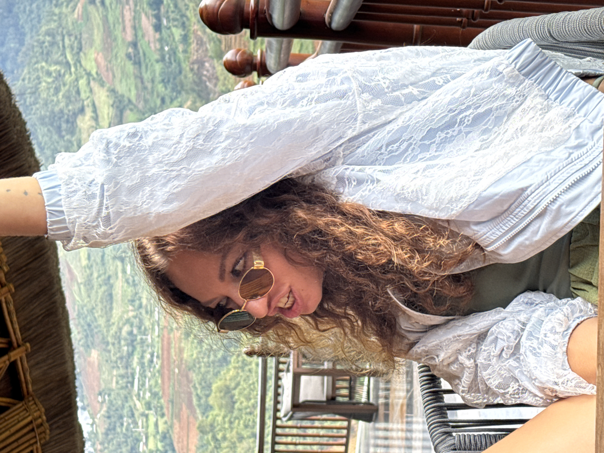 This vibrant outdoor portrait captures a young woman smiling broadly against a picturesque mountainous backdrop in Púng Luông, Vietnam. The image shows the woman positioned with her head towards the right side of the frame, looking towards the left, with her body angled and one arm raised and extended towards the upper left. She has long, voluminous curly brown hair and wears stylish round, gold-rimmed sunglasses. Her wide, joyful smile reveals her teeth, conveying a sense of happiness and relaxation. She is dressed in a light blue or white lace bomber-style jacket, layered over what appears to be a dark green top.
The setting is an elevated vantage point, likely a balcony or terrace, offering expansive views of the lush green landscape. In the background, rolling mountains are visible, characterized by terraced fields or agricultural plots that mark their slopes, indicative of the region's geography. A rustic thatched roof structure is partially visible on the far left, hinting at local architectural elements. Wooden railings, possibly part of a balcony or outdoor seating area, are also discernible. The clear visibility and the presence of sunglasses suggest a sunny and pleasant daytime environment. The overall impression is one of enjoyment and appreciation for the beautiful natural surroundings. No visible text is present in the media file.