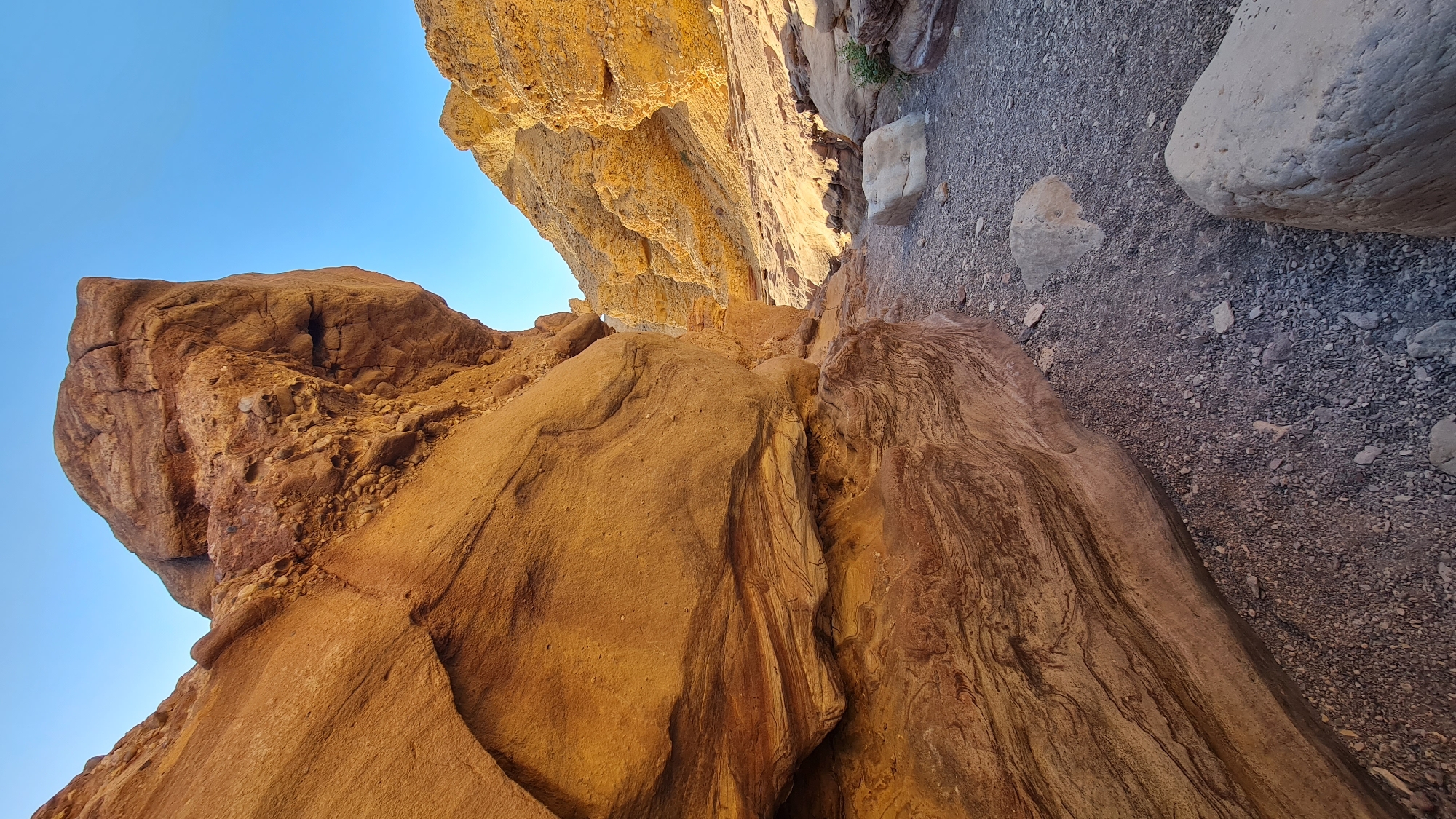 The image depicts a desert canyon landscape in Hevel Eilot Regional Council, Israel. The scene is dominated by large, ochre-colored rock formations with visible stratification. A gravel path runs along the canyon floor. The sky is a clear, bright blue. The rock formations are weathered and eroded. The sunlight is warm and casts shadows.