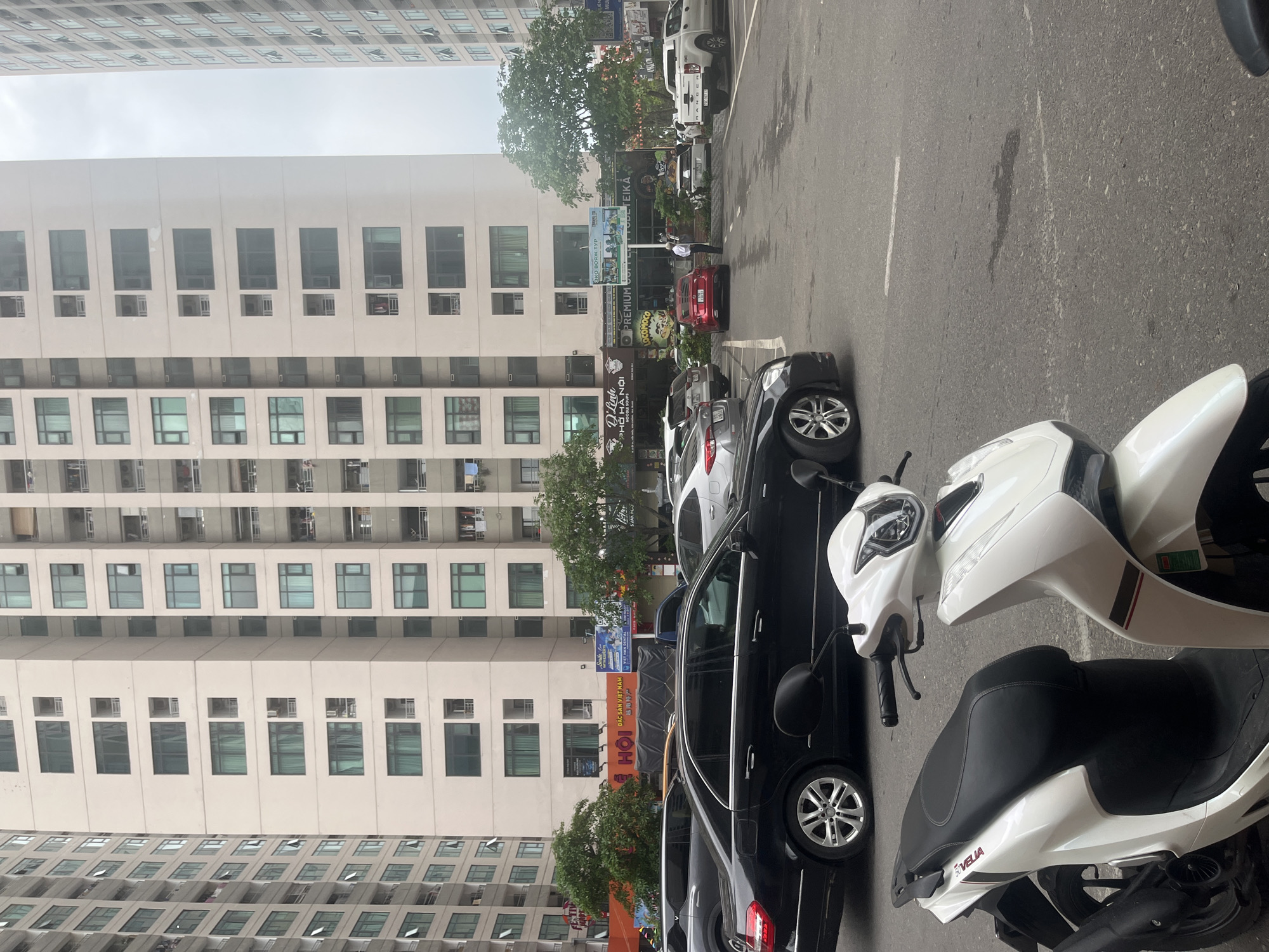 This image captures an urban street scene in Bac Nha Trang Ward, Vietnam, under an overcast sky during daytime. The scene is dominated by tall, multi-story buildings with numerous rectangular windows, indicative of a residential or mixed-use complex, lining the left side of the frame.
In the foreground and mid-ground, a variety of vehicles are parked along a paved street or parking area. A white scooter, branded "SOVELIA," is prominently featured in the lower right, with its handlebars and front visible. Behind it, several cars are parked, including a black sedan closest to the viewer, a silver car, a red car, and a white pickup truck (possibly a Ford Ranger). One person, dressed in a white shirt, is visible walking on the street near the parked vehicles.
The lower levels of the buildings house commercial establishments, with several signs visible. Notable text includes "HỘI," "ĐẶC SẢN VIỆT NAM" (Specialties of Vietnam), "D'Linh PHỞ HÀ NỘI NOODLE SOUPS," "VIET HAN DENTAL," "SHAN FOO," "PREMIUM," "LOCOMOCO," and "КЭКО ВОЯЖ ТУР." The overcast weather creates soft, even lighting without strong shadows. The street surface is dark asphalt with faint white markings. The overall impression is a bustling urban environment typical of a Vietnamese city.