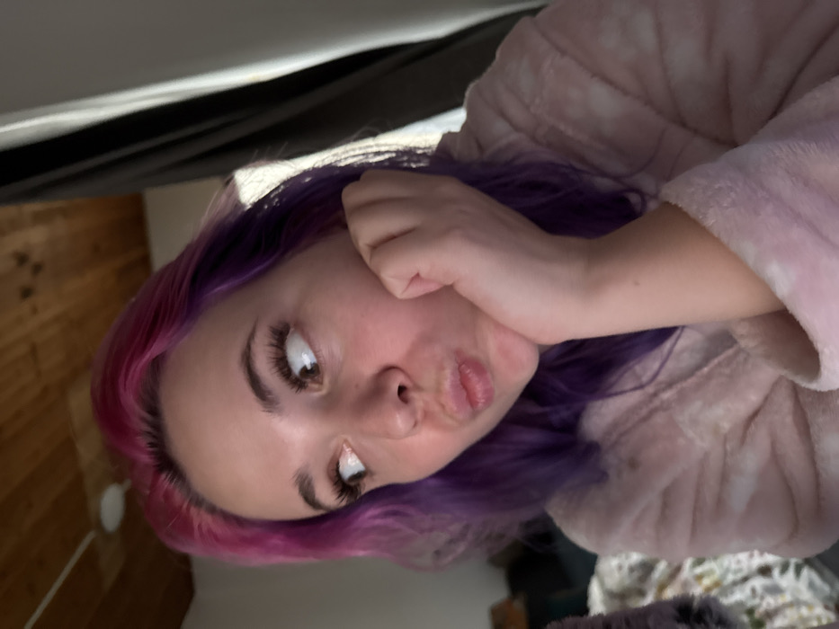 A woman with purple hair and pink roots poses with pursed lips and a hand against her cheek. She gazes toward the camera with large, emphasized eyelashes. The woman is wearing a fuzzy pink robe. She appears to be positioned underneath something dark and possibly fabric. A wooden panel wall is visible in the background.