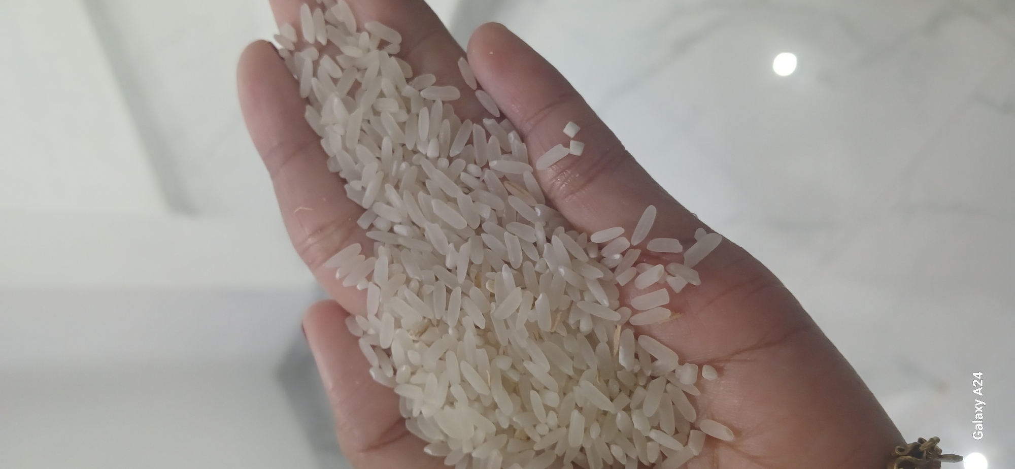 A close-up, high-angle shot shows a person's palm filled with white rice grains. The palm is cupped, holding a substantial amount of uncooked rice. The background is out of focus, appearing as a light-colored, possibly marbled surface. The lighting suggests an indoor setting. The image is geotagged to Kuwarisan, Indonesia.