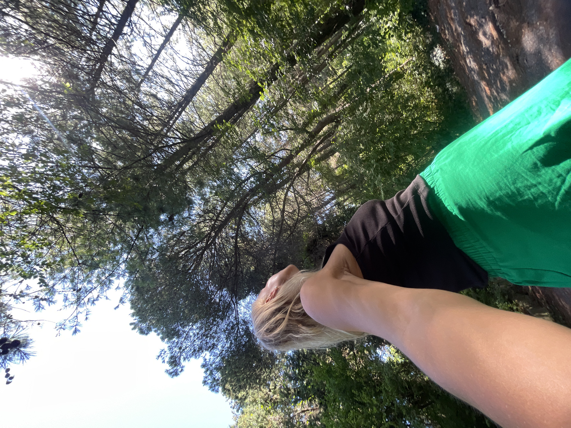 An overhead, wide-angle shot captures a blonde woman in a black tank top and bright green pants, looking up at the sky as she stands in a forest. The woman is positioned on the left side of the frame, with her arm extending towards the viewer, obscuring part of the lower left quadrant of the image. Her hair is windswept, and her face is partially visible from the side, tilted upwards. The forest surrounds her, with tall trees and lush green foliage filling the background. Sunlight filters through the canopy, creating dappled light on the forest floor. The ground appears to be a mixture of dirt and fallen leaves. The image does not contain any elements that definitively place it in Sochi, Russia.